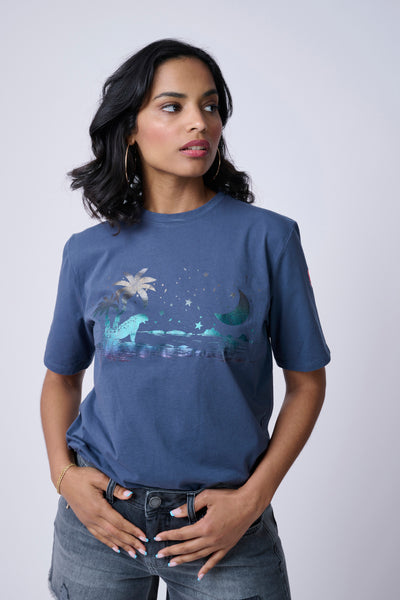 Scamp and Dude Blue T-Shirt with Palm Beach Graphic on Front | Model wearing a blue top with beach design on front with grey jeans