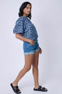 A woman wearing a soft blue with black floral leopard and lightning bolt print pintuck sleeve T-shirt with blue denim shorts