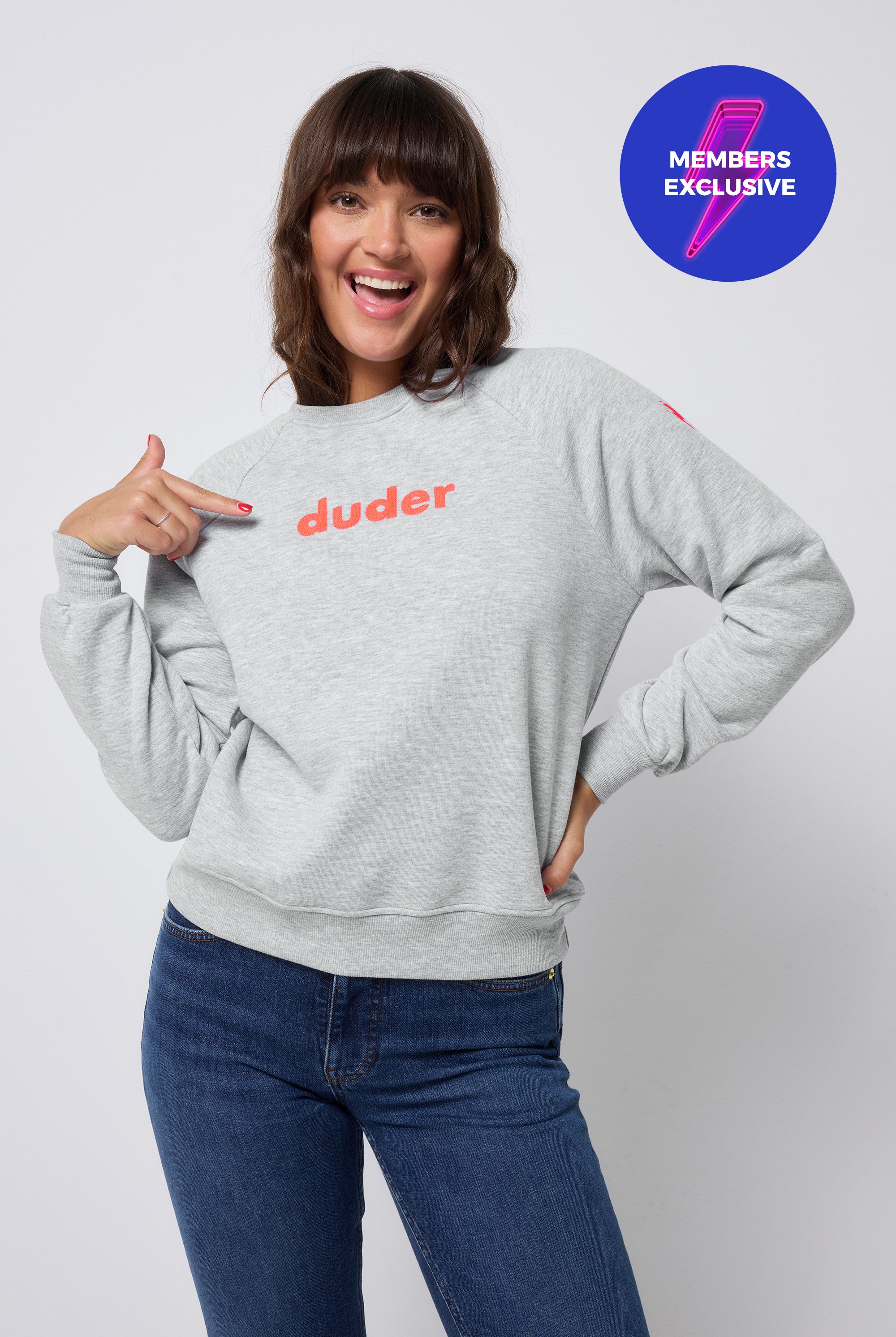 Scamp and Dude Grey Marl Duder Embroidery Oversized Sweatshirt | Model with dark hair wearing a grey crew neck sweatshirt with dark blue jeans. Sweatshirt features the word 'duder' embroidered on the chest in a neon coral thread.
