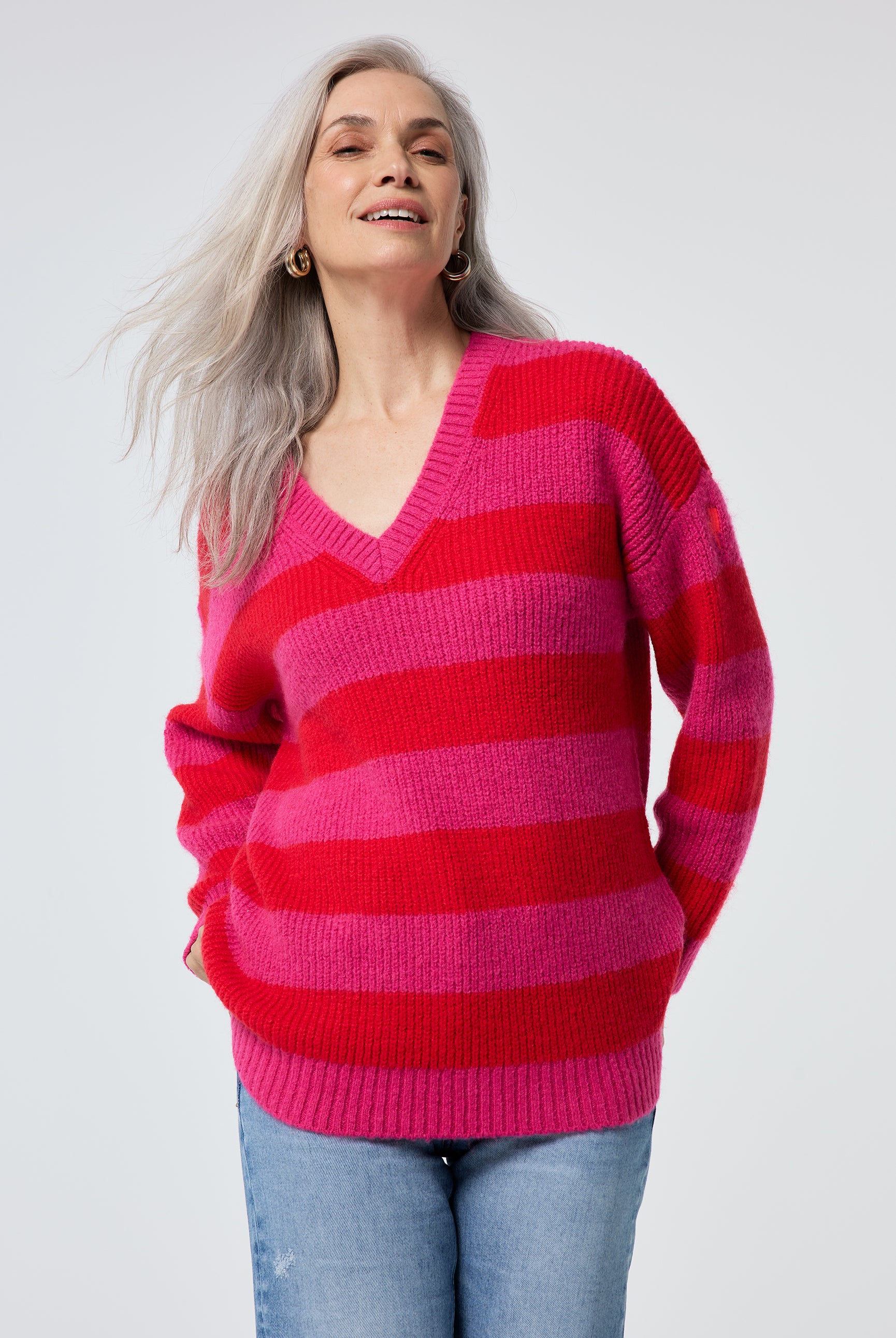 Scamp and Dude Pink and Red Stripe V Neck Knit Jumper | Model wearing a long sleeve v neck knit jumper with pink and red stripes paired with distressed blue jeans.