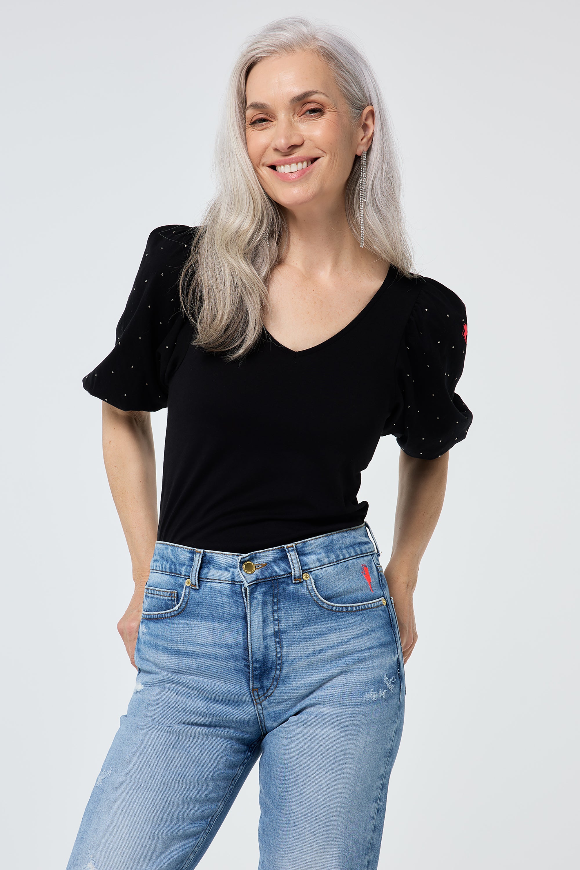 Scamp and Dude Black Studded V-Neck Puff Sleeve T-Shirt | Model wearing a black v neck t shirt featuring puff sleeve with stud detail, paired with distressed light blue jeans and heels.