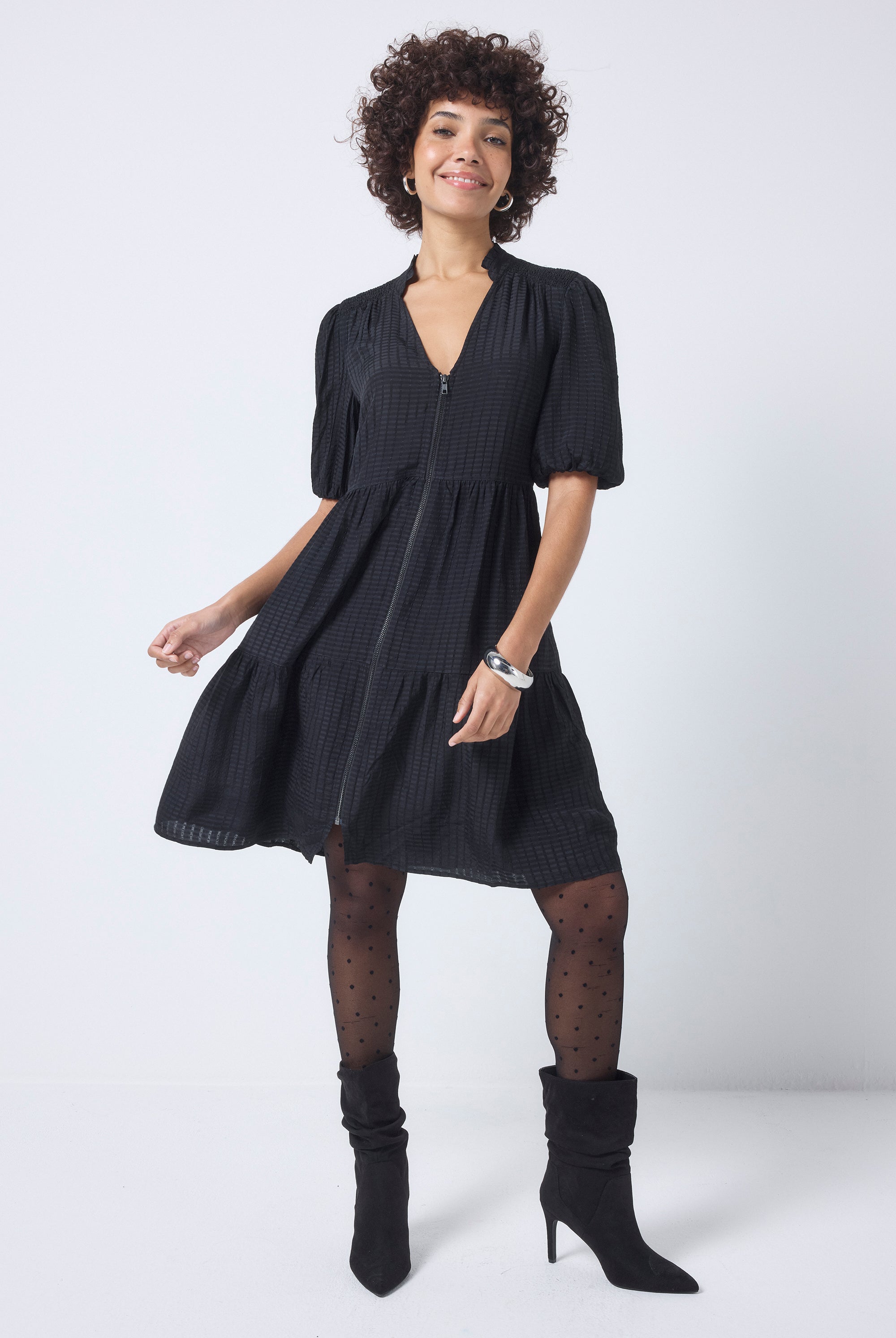 Scamp and Dude Black Textured Zip Detail Tiered Short Dress | Model wearing a short black textured dress with a zip front detail, worn with black polka dot tights and boots.