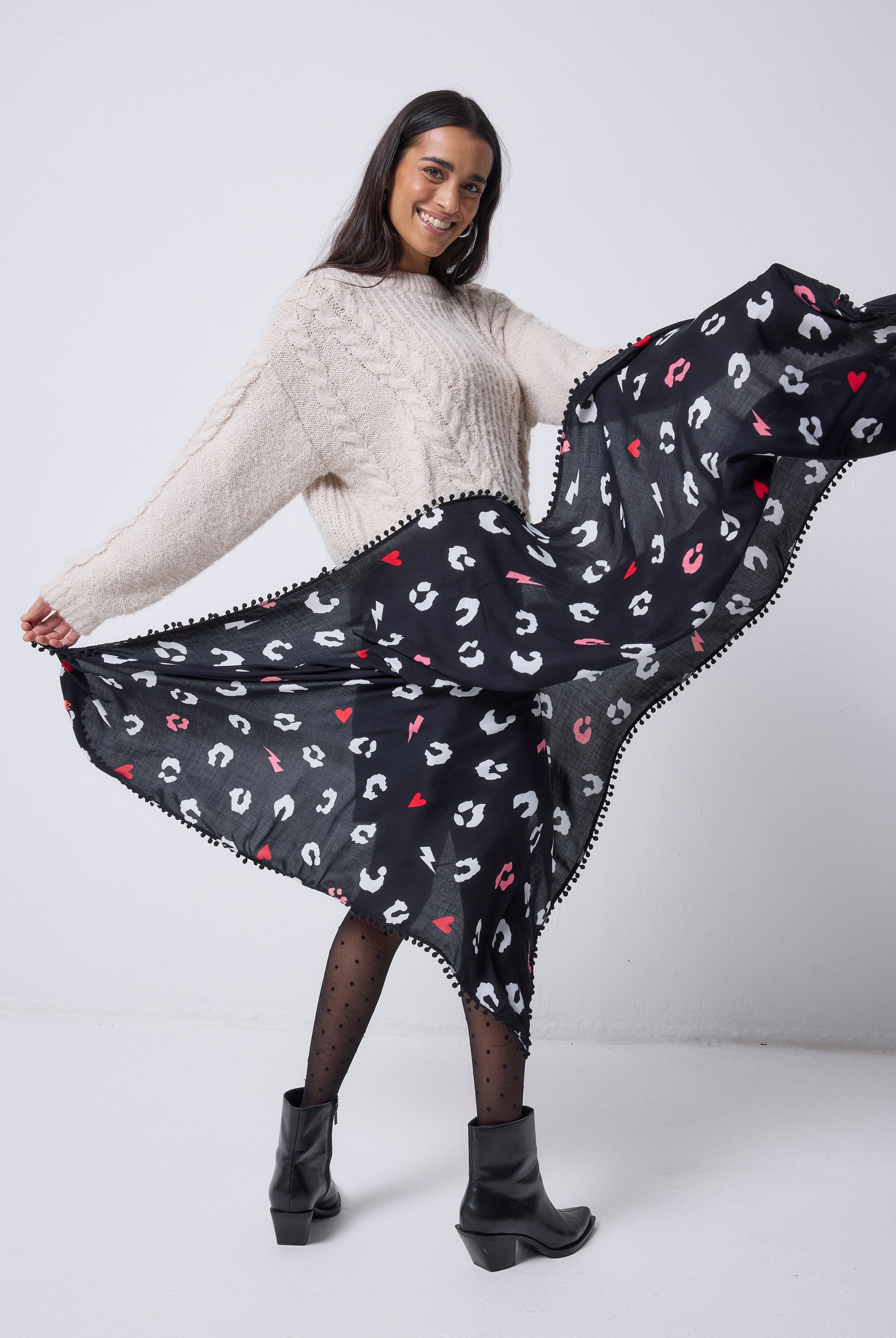 Scamp and Dude Black with White Love Leopard Charity Super Scarf | Model wearing an oatmeal cable knit jumper with a black denim skirt, tights and boots. Holding a black scarf with white love leopard print.