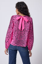 Scamp and Dude Pink with Black Wild Leopard Blouson Sleeve Top | Model with dark curly hair is wearing a blue pair of jeans with a pink leopard print blouse. The blouse has a tie back feature which has been tied into a bow.