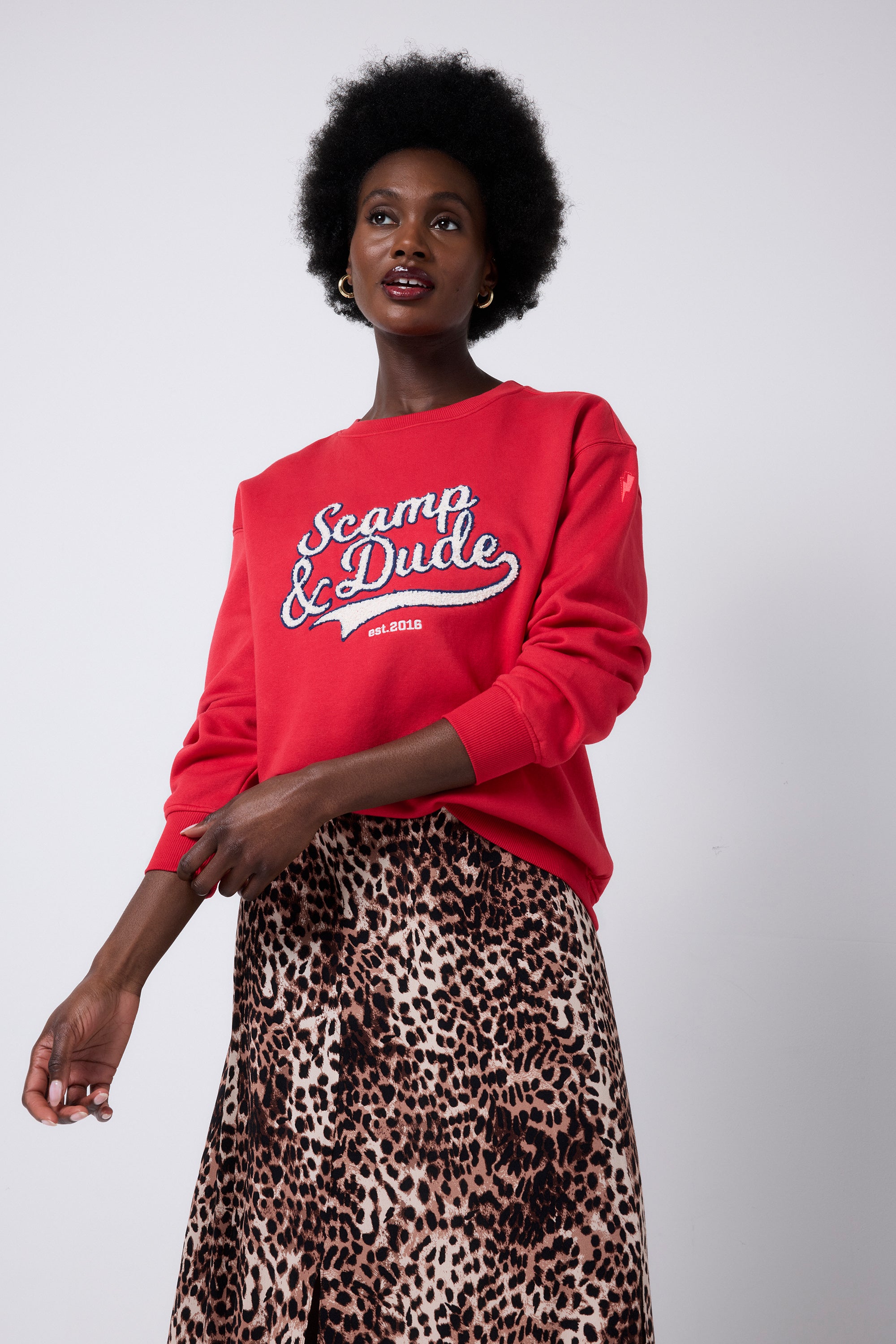 Scamp and Dude Red Varsity Scamp & Dude Oversized Sweatshirt | Model wearing a neutral cheetah print skirt with an oversized red crew neck sweatshirt with Scamp & Dude logo.