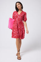 Scamp and Dude Pink and Red Palm Zip Detail Tiered Short Dress | Model wearing a short sleeve pink mini dress with red palm print all over. The dress has a zip feature on the front. Worn with a pink bag and brown sandals.