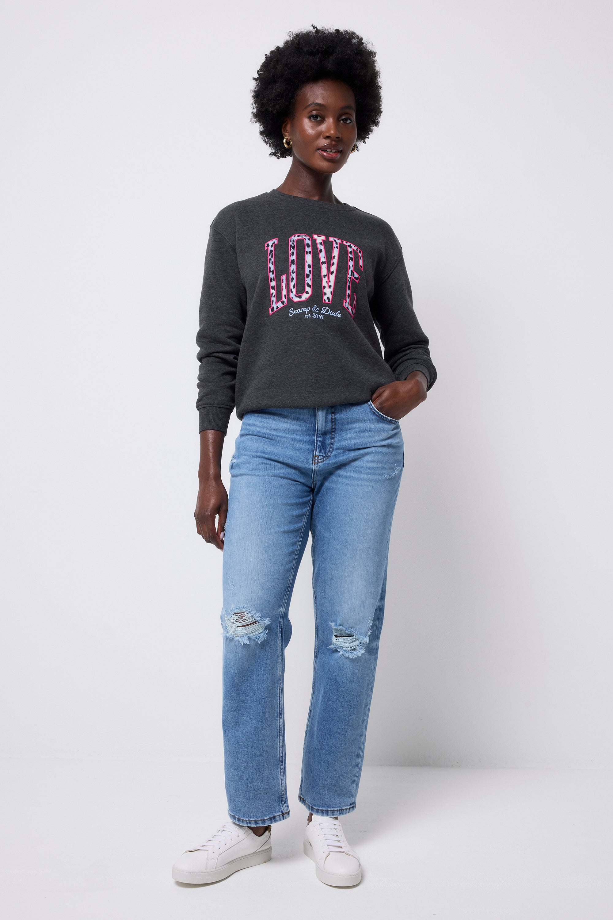 Scamp and Dude Dark Grey Love Cheetah Oversized Sweatshirt | Model wearing blue distressed jeans with a dark grey oversized sweatshirt. The sweatshirt has 'LOVE'  in a blue and white cheetah print across the front of the sweatshirt with pink embroidery.
