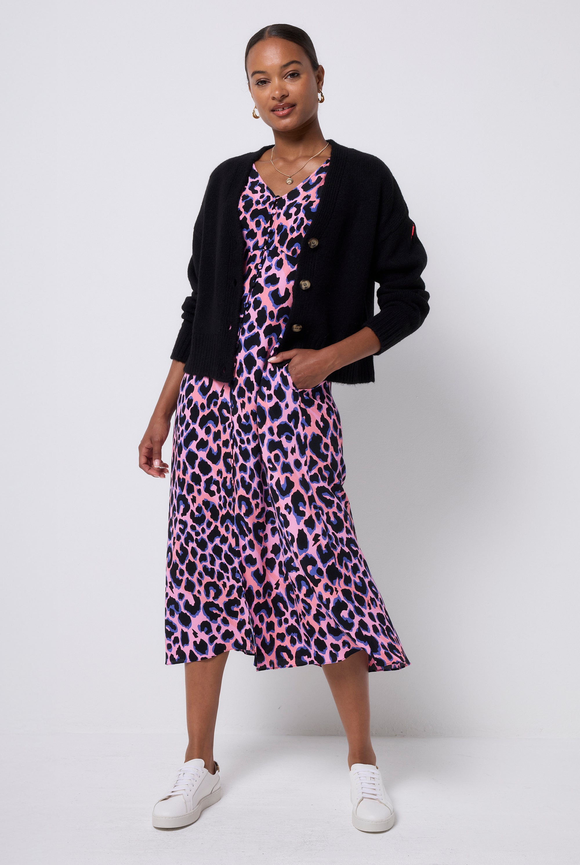 Scamp and Dude Black Cardigan | Model wearing a pink with blue and black leopard print dress with a black cardigan worn over it. Paired with white trainers and gold jewellery.