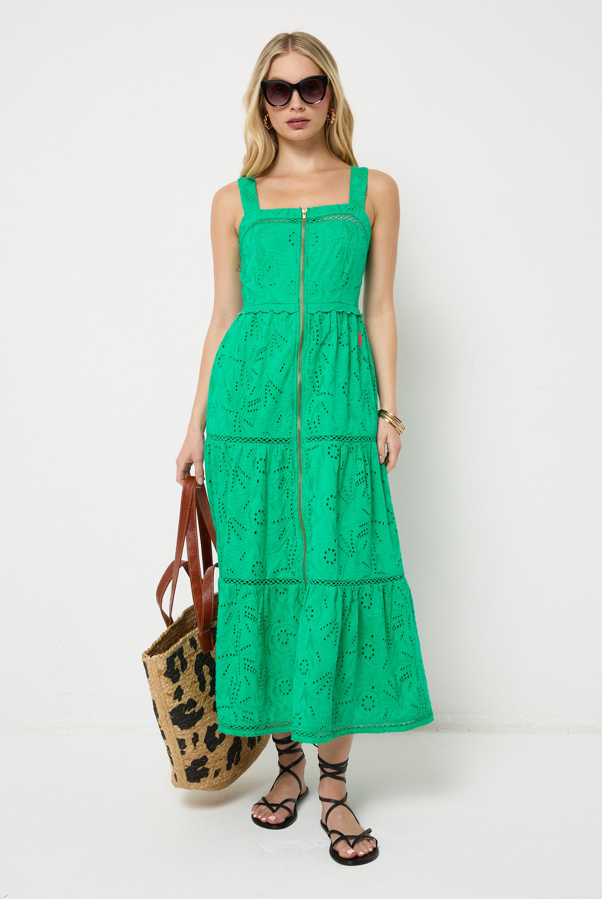 Scamp and Dude Green Broderie Zip Detail Midi Sundress | Model with blonde hair wearing a green broderie zip front midi dress with black sandals and sunglasses, holding a leopard print straw bag.