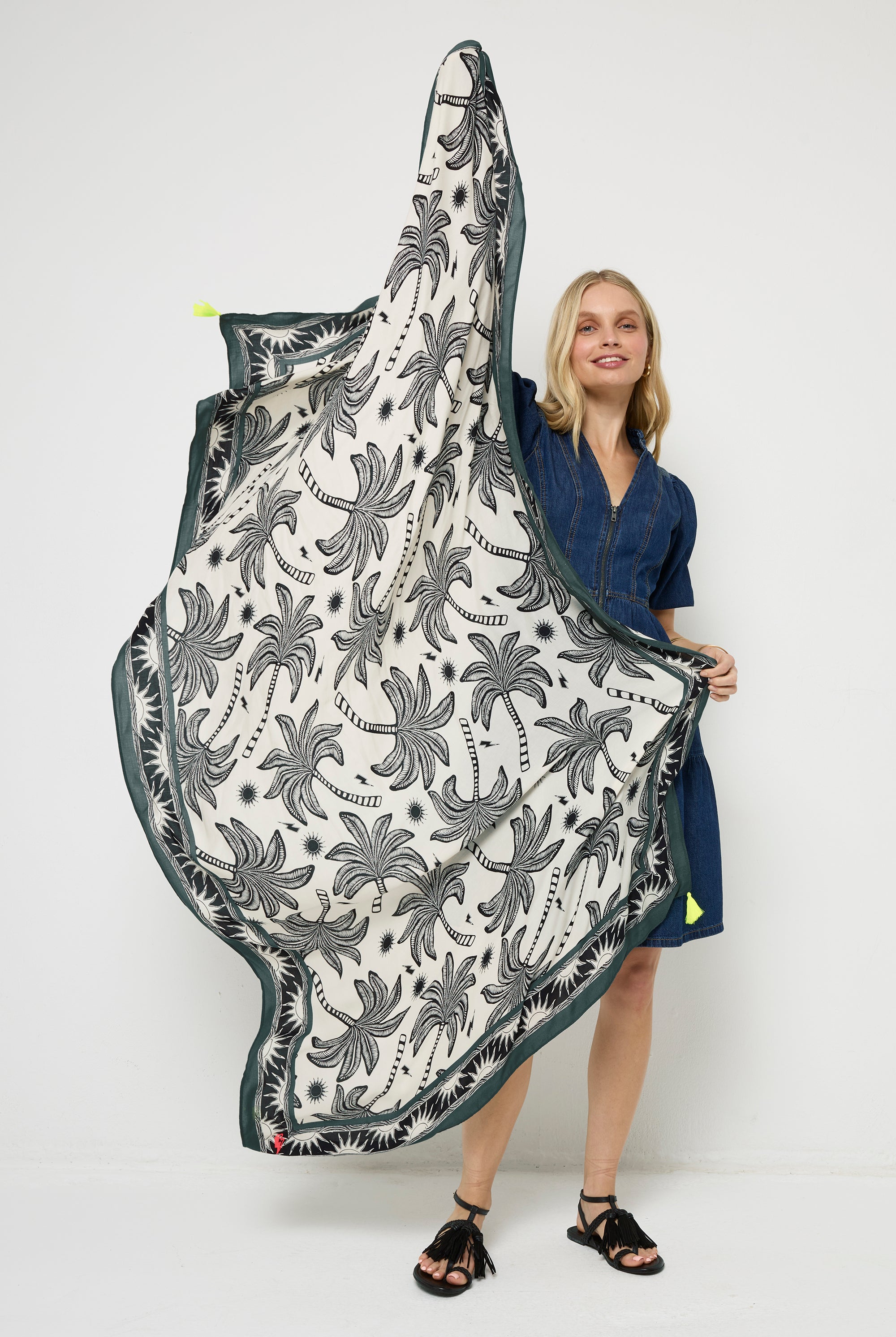 Scamp and Dude White with Black and Khaki Palm Spring Charity Super Scarf | Model wearing a short dark blue denim dress with black sandals, holding a white and khaki palm print super scarf.