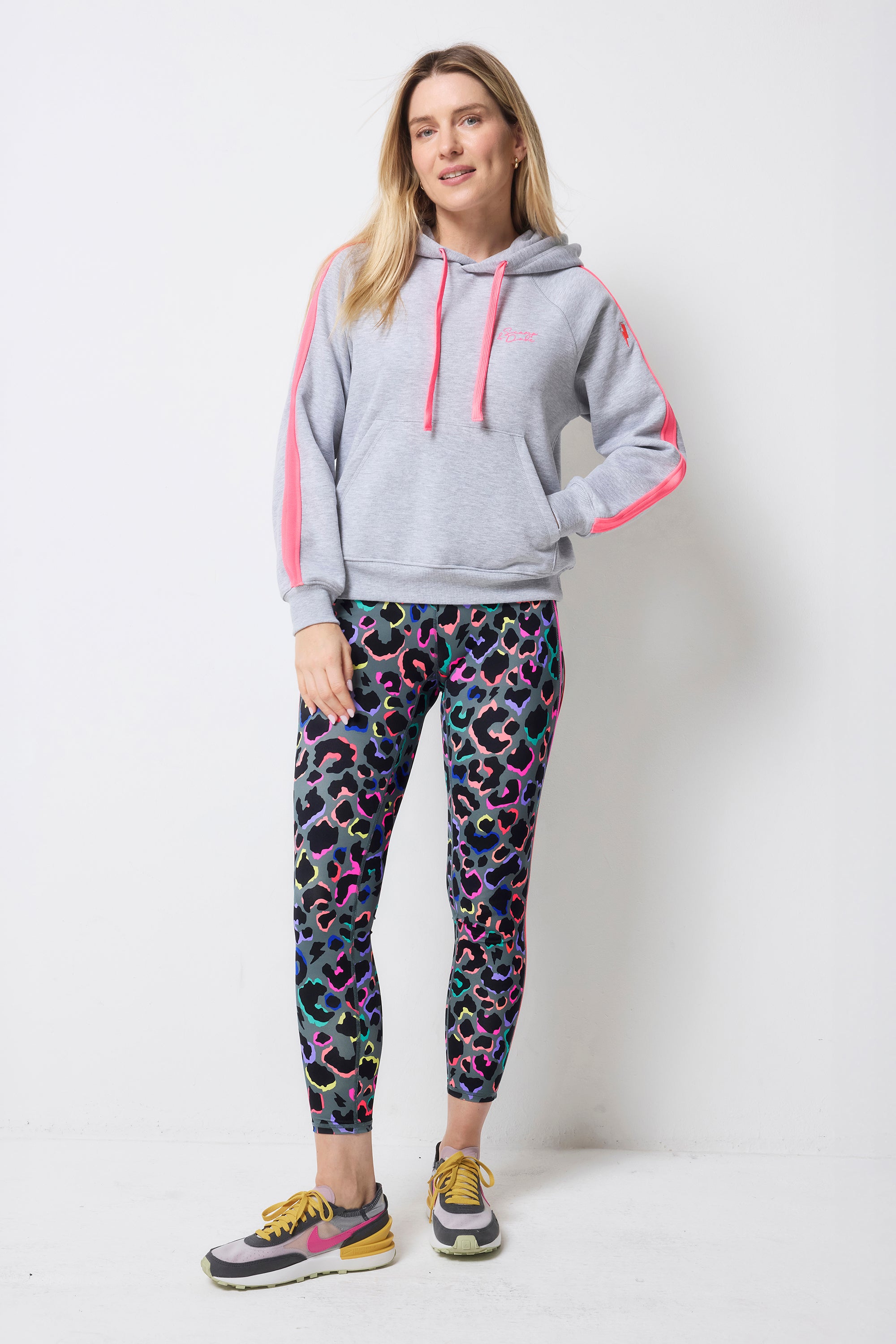 Scamp and Dude Dark Khaki with Rainbow Shadow Leopard 7/8 Length Active Leggings | Model wearing a grey hoodie with neon details paired with a pair of khaki 7/8 leggings with rainbow leopard print and white colourful trainers.