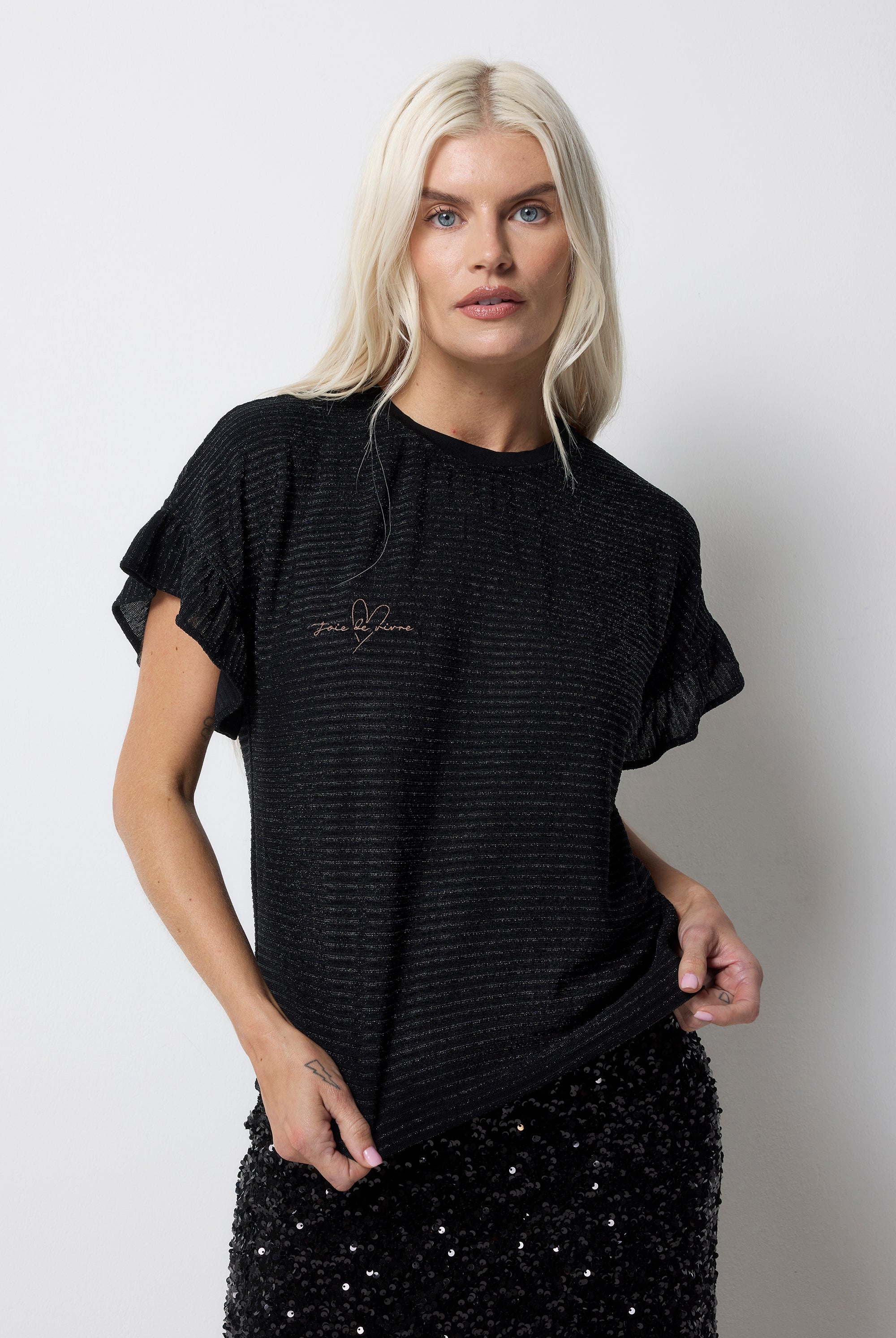 Scamp and Dude Black Lurex Frill Sleeve T-Shirt | Model wearing a black lurex stripe t-shirt with frill sleeves and embroidered slogan. Paired with a black sequin midi skirt.