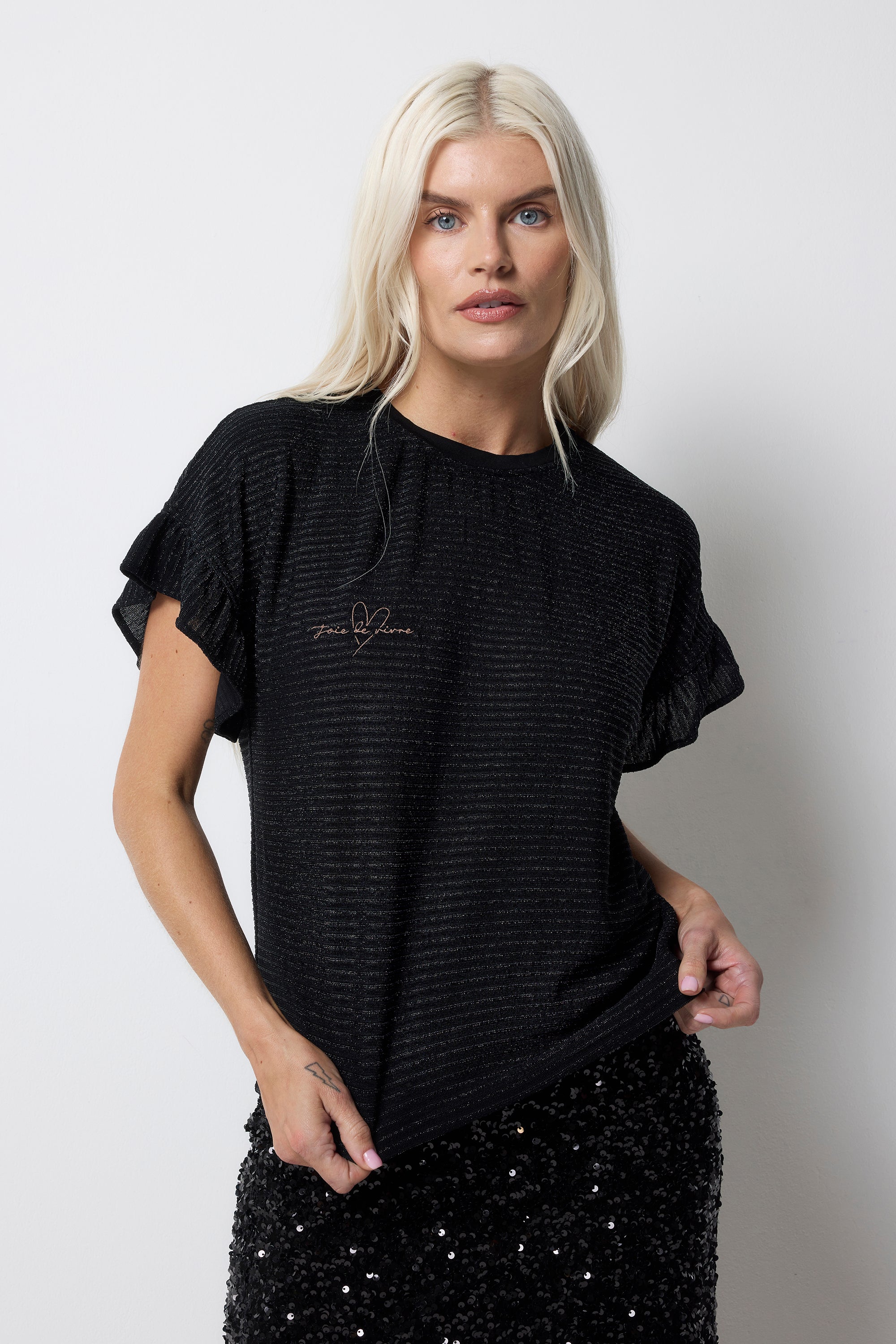 Scamp and Dude Black Lurex Frill Sleeve T-Shirt | Model wearing a black lurex stripe t-shirt with frill sleeves and embroidered slogan. Paired with a black sequin midi skirt.