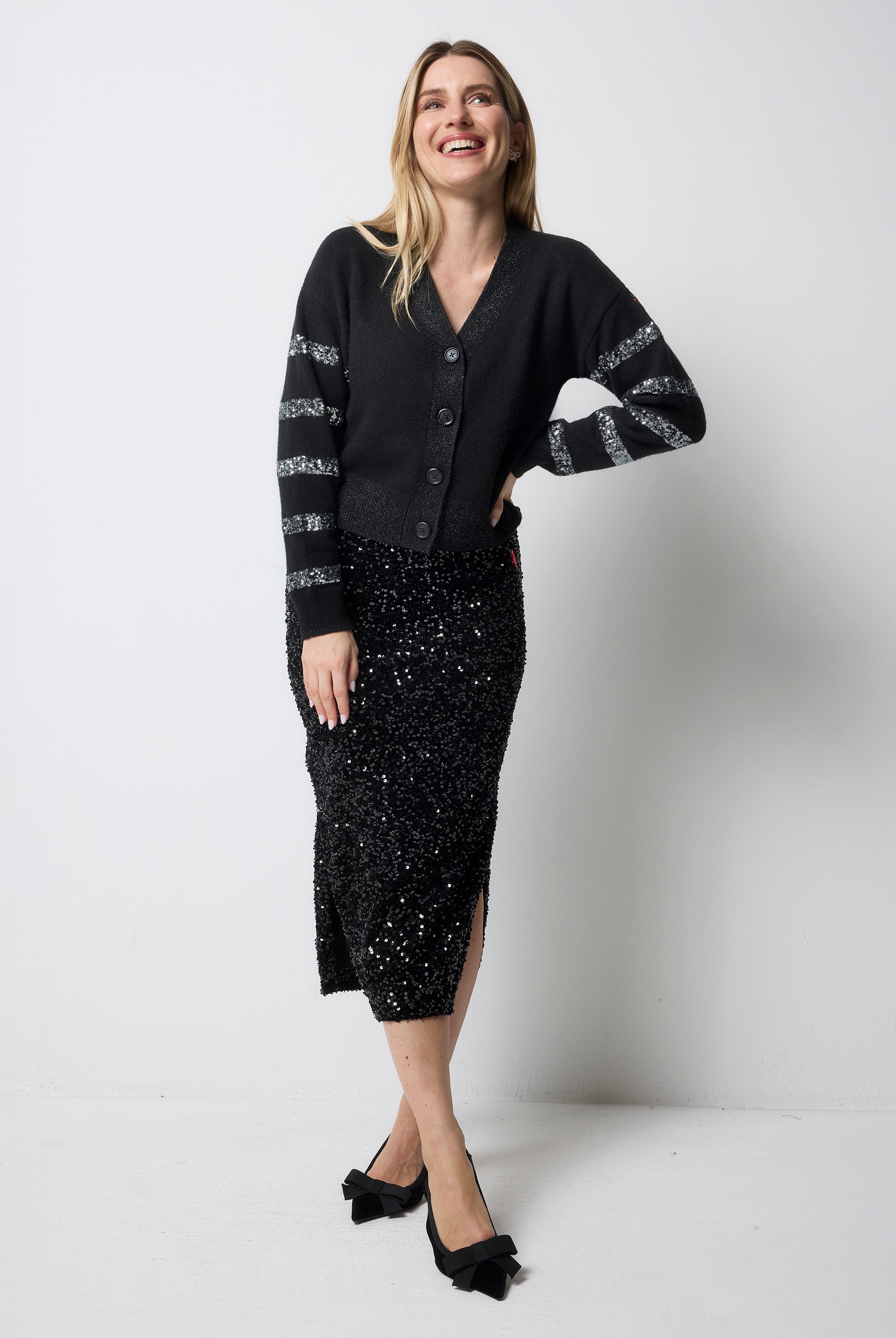 Scamp and Dude Black with Pewter Sequin Stripe Cardigan | Model wearing a black cardigan with sequin sleeves and a black sequin skirt with black bow heels.