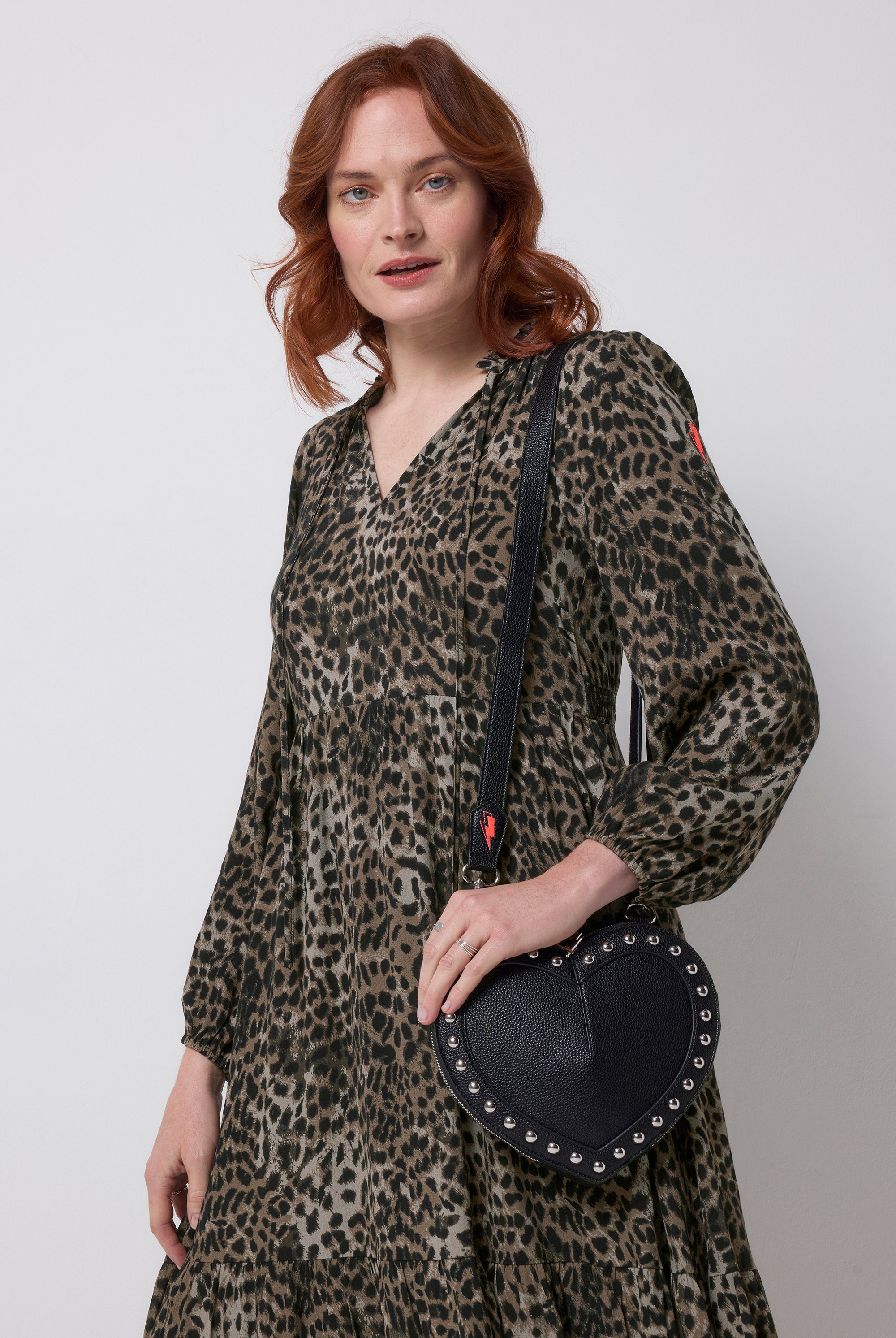 Scamp and Dude Black Heart Studded Cross Body Bag | Model wearing a khaki leopard print long sleeve dress with a black heart shaped bag with silver stud detail.