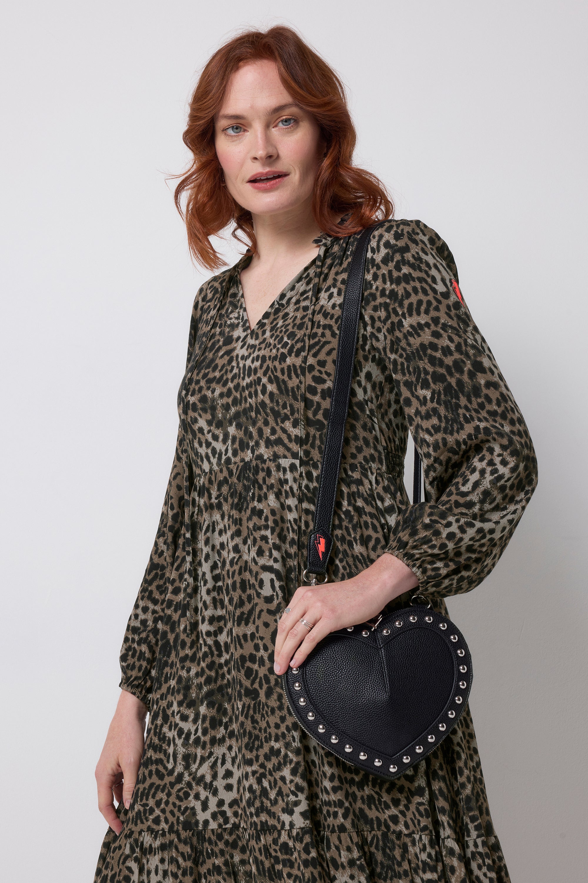 Scamp and Dude Black Heart Studded Cross Body Bag | Model wearing a khaki leopard print long sleeve dress with a black heart shaped bag with silver stud detail.