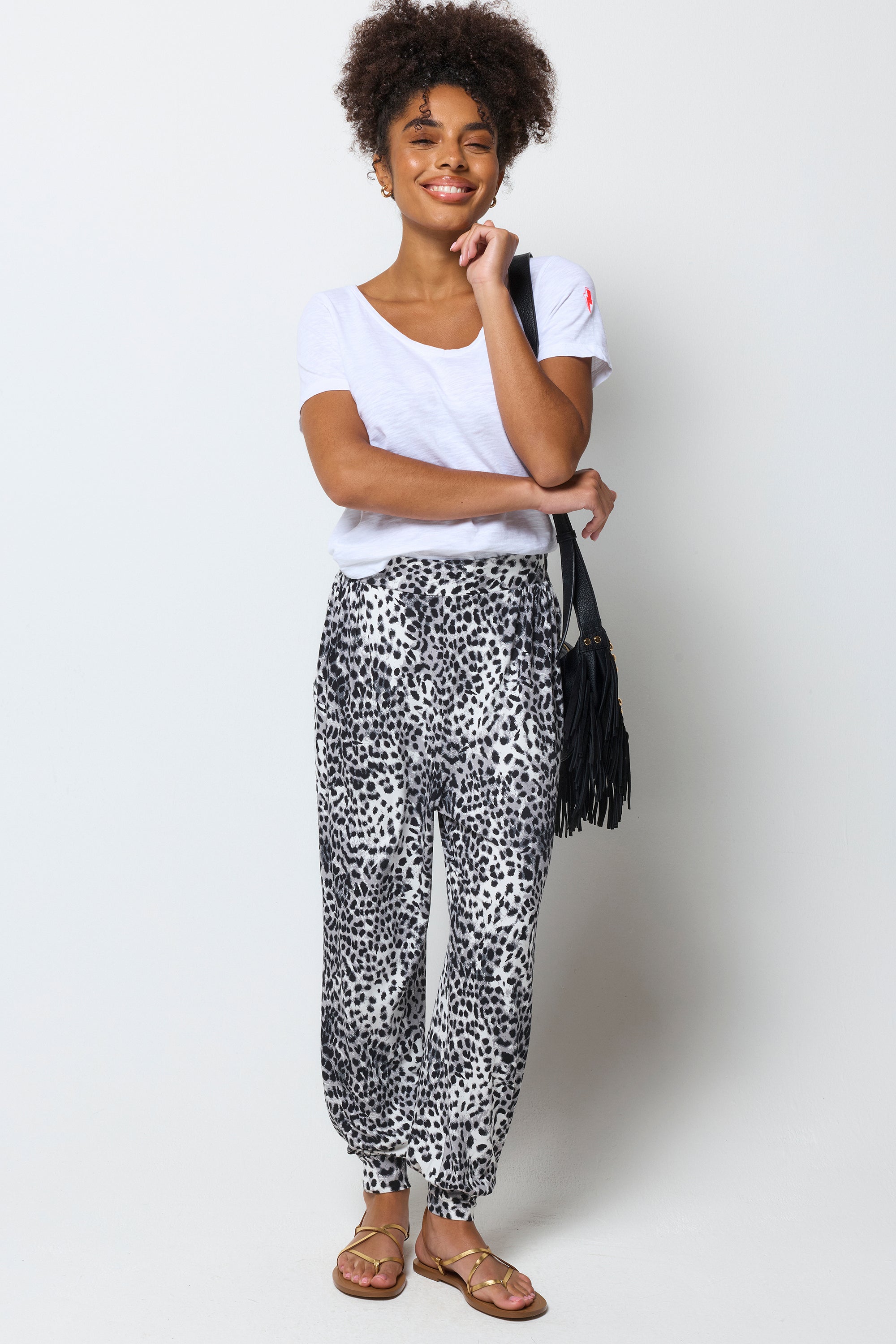 Scamp & Dude Grey Cheetah Balloon Leg Jersey Trouser | Model wearing a white v-neck t-shirt with a pair of balloon leg jersey trousers in a grey cheetah print. Worn with gold sandals and a black fringe bag.