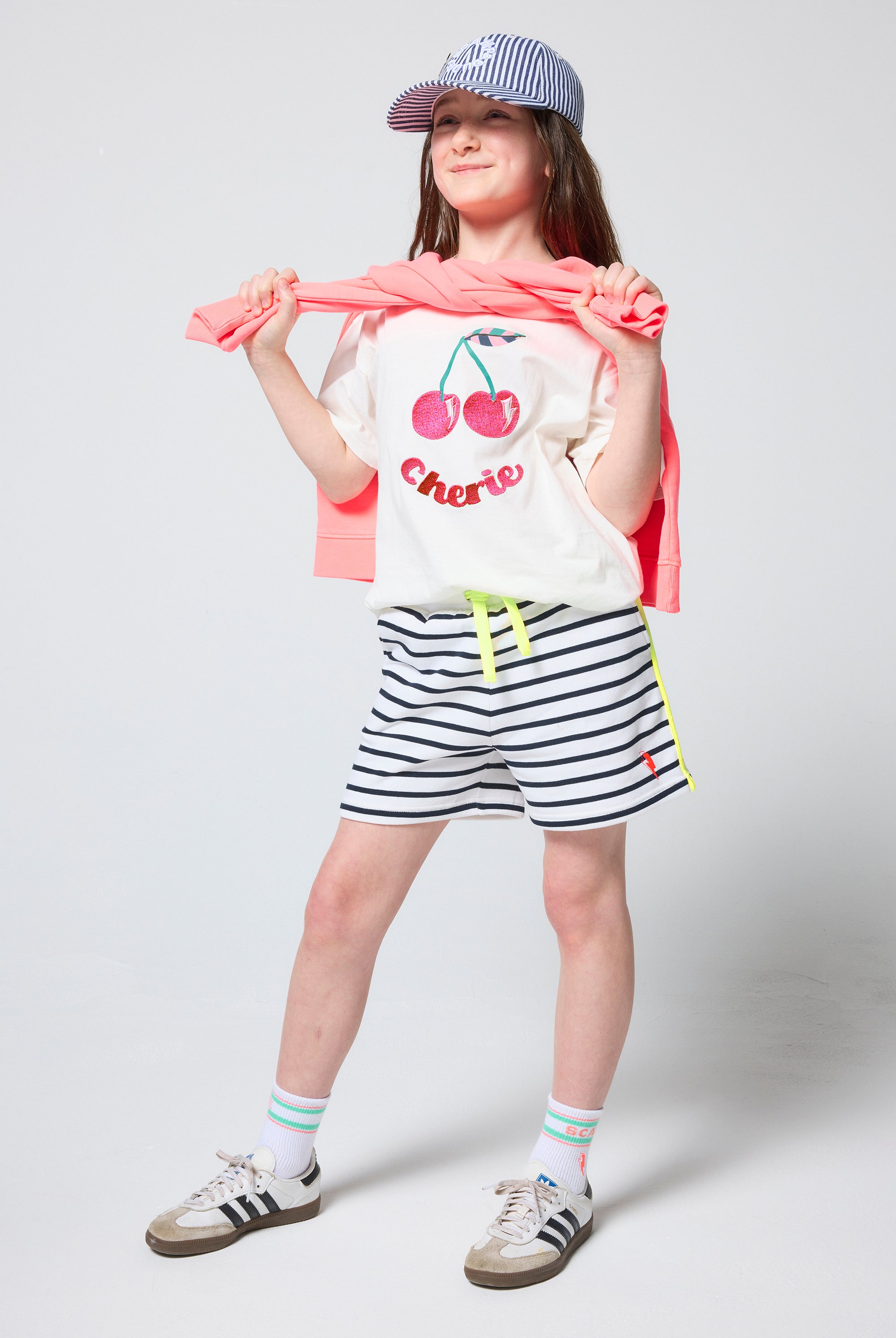 Scamp & Dude Kids White Cherry Print T-Shirt | Model wearing a cherry-print shirt, striped shorts, and a cap.