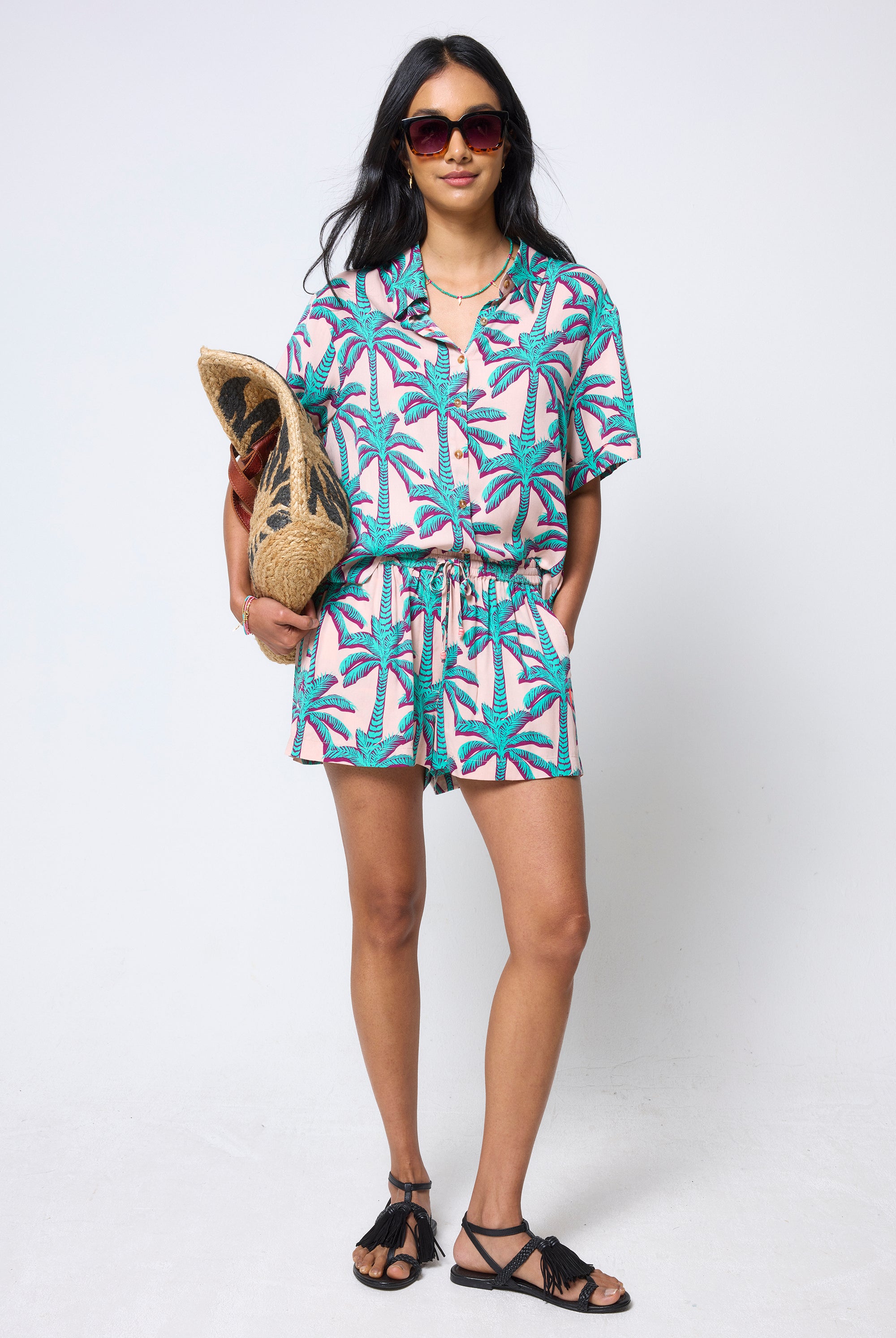 Scamp & Dude Cream with Green Shadow Palm Shirt | Model wearing a palm print shirt and shorts holding a straw bag against a white background.