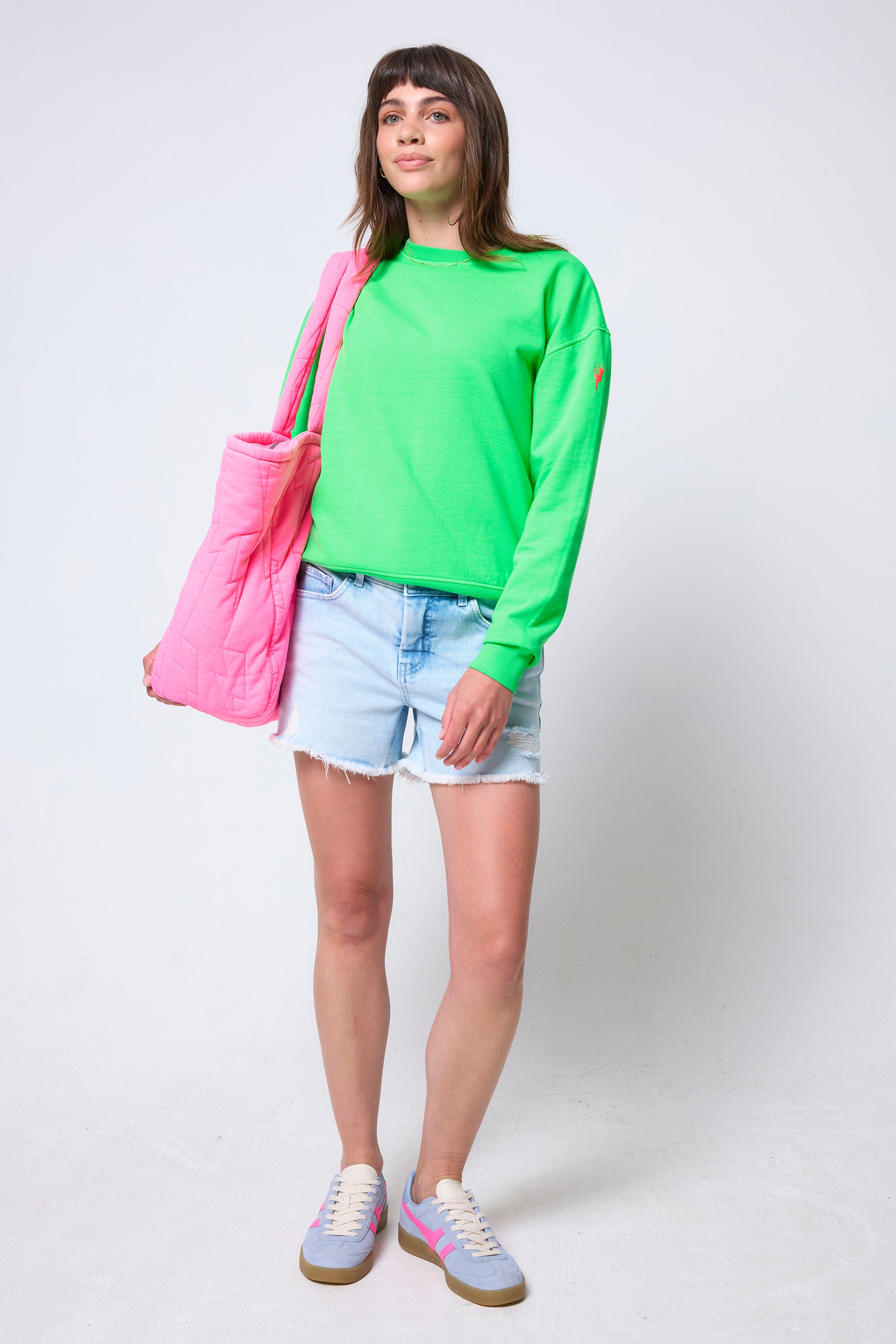 Scamp & Dude Neon Green Washed Ultimate Sweatshirt | Model wearing a bright green sweatshirt, denim shorts, and blue trainers, holding a pink bag.