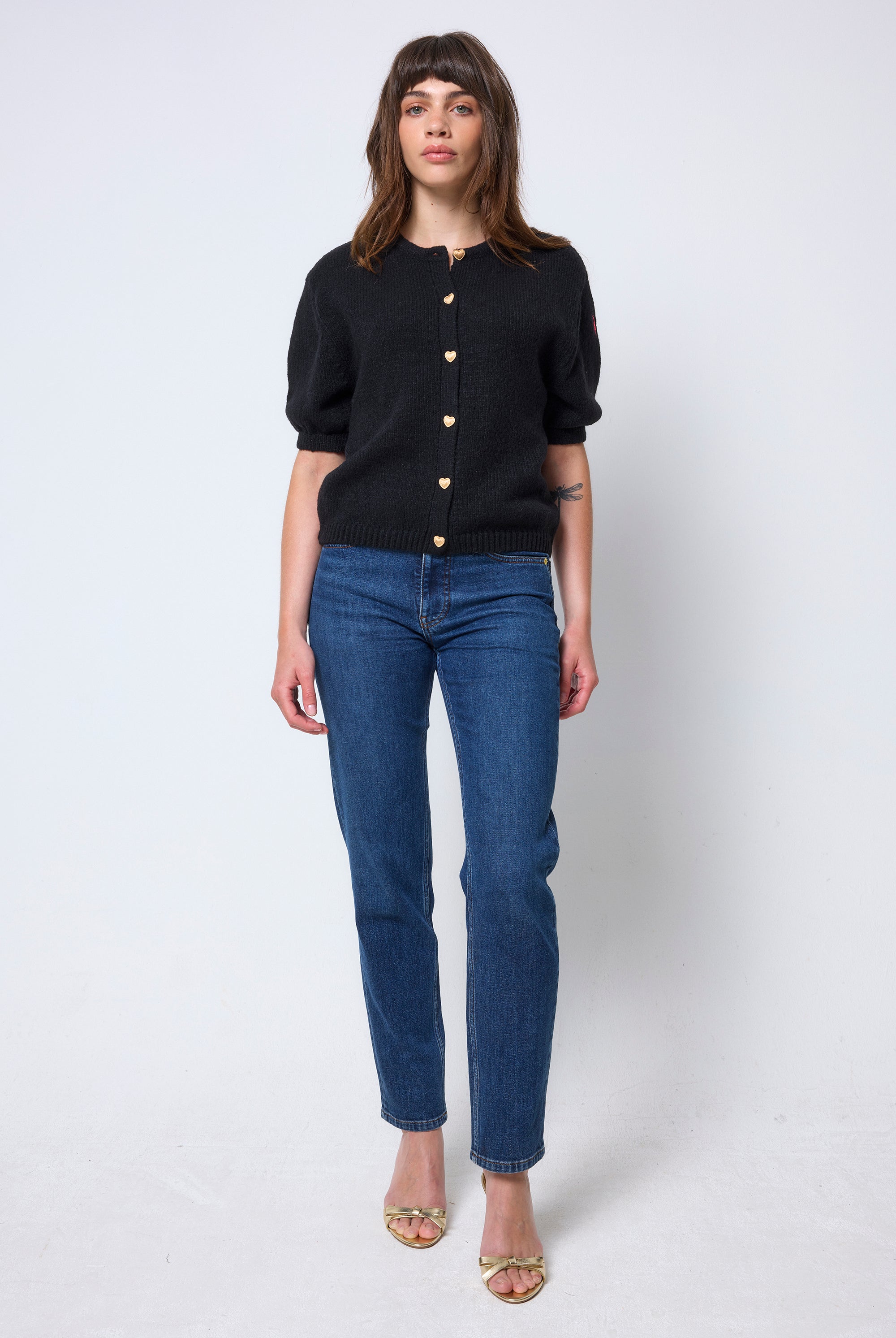 Scamp & Dude Black Short Sleeve Cardigan | Model wearing a black short sleeve cardigan with gold heart button detail. Worn with blue boyfriend jeans and heels.