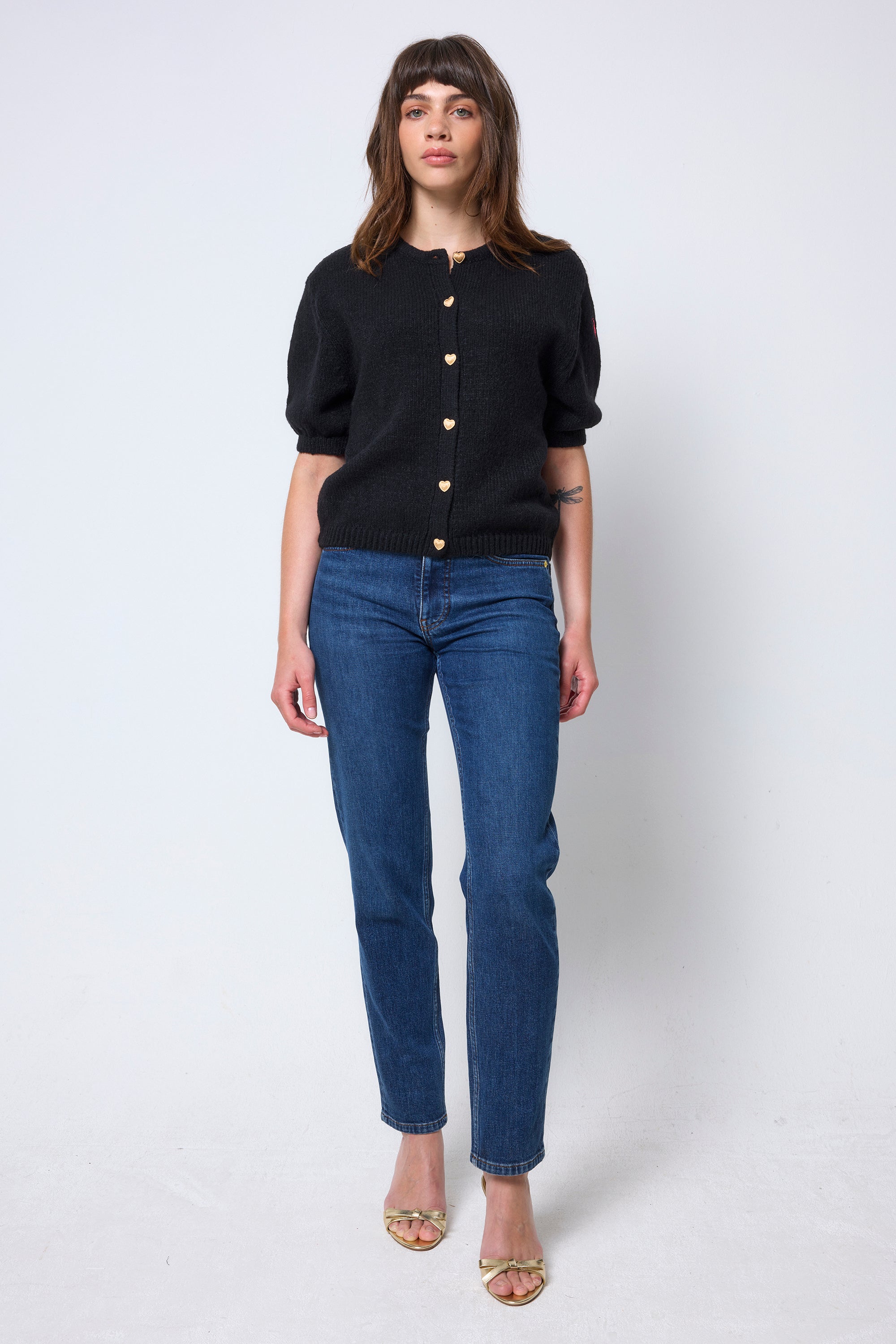 Scamp & Dude Black Short Sleeve Cardigan | Model wearing a black short sleeve cardigan with gold heart button detail. Worn with blue boyfriend jeans and heels.