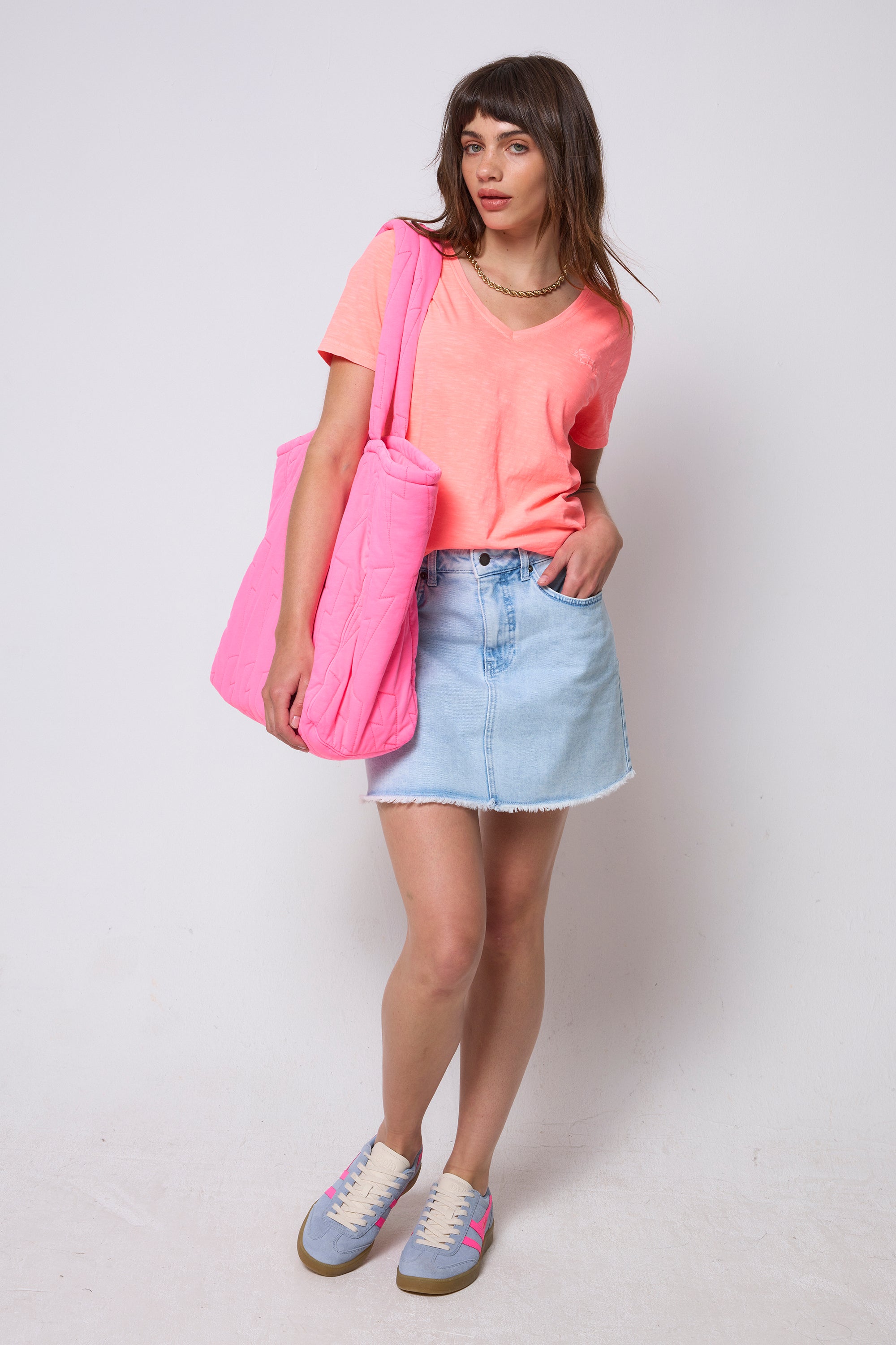 Scamp & Dude Washed Neon Coral V-Neck T-Shirt | Model wearing a bright coral v-neck t-shirt with a blue denim skirt and blue trainers. Holding a neon pink quilted tote bag.