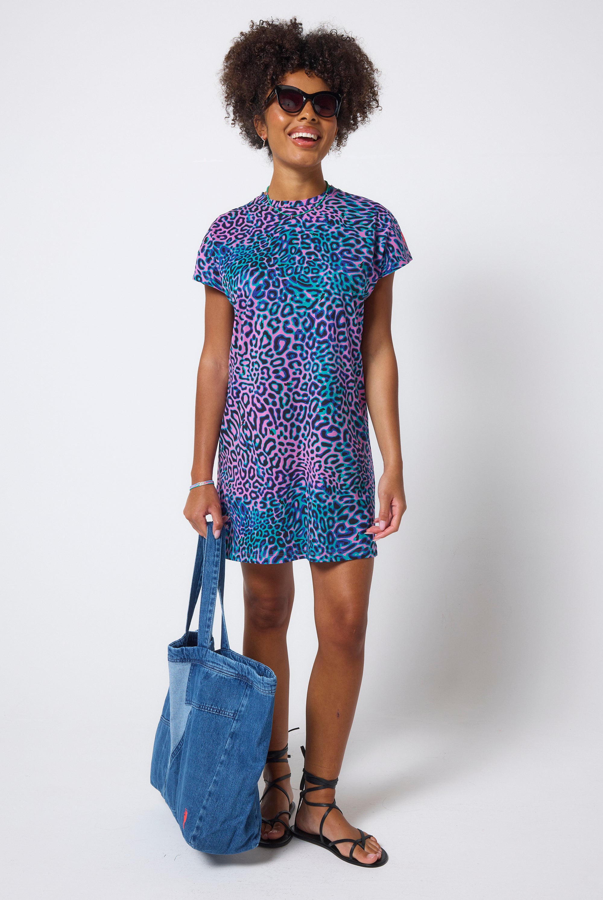 Scamp & Dude Pink with Green Leopard Ray T-Shirt Dress | Model wears a short t-shirt dress in a pink and green leopard ray print. Worn with black strappy sandals and a denim tote bag.