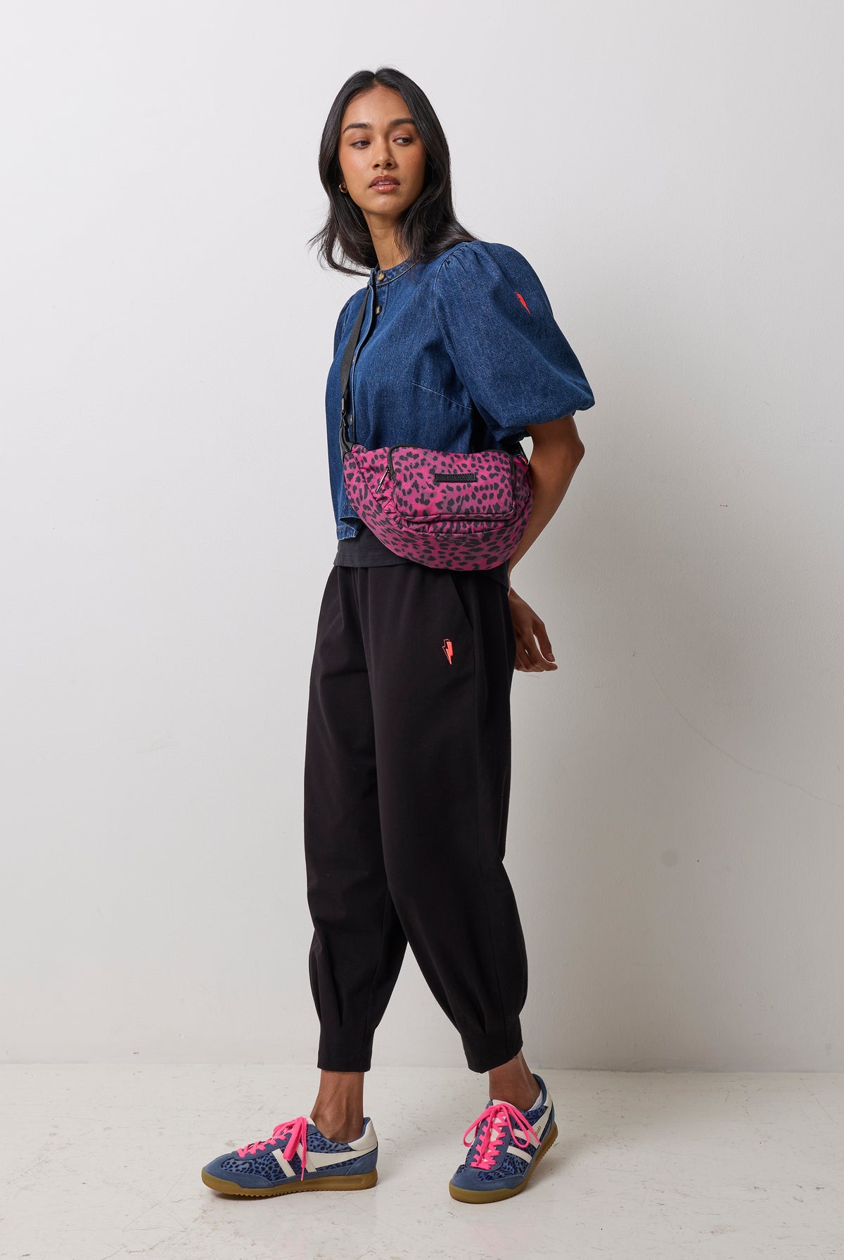 Scamp & Dude Black Cherry Cheetah Bum Bag | Model wearing a blue cropped jacket with puff sleeves, black jogger pants, and blue sneakers with bright pink laces. She is carrying a pink and black leopard-print bum bag.