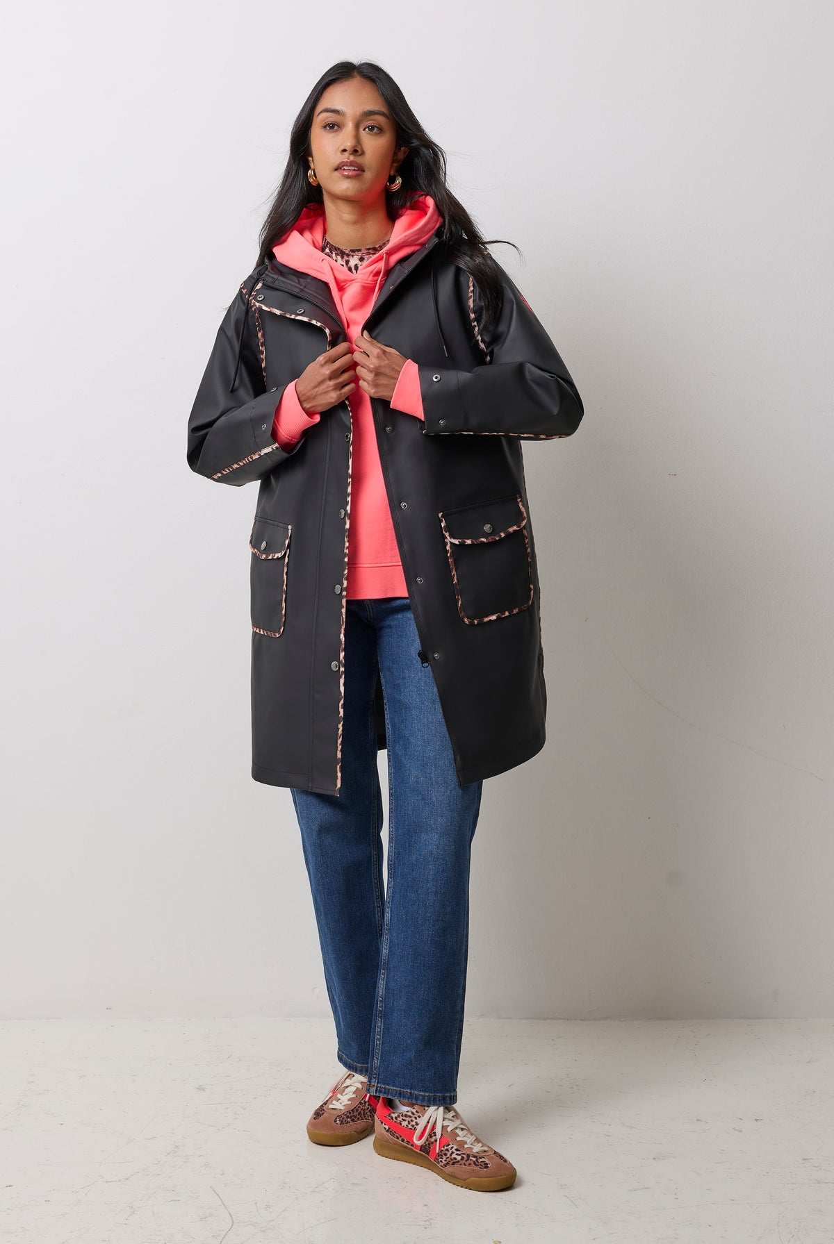 Scamp & Dude Black with Neutral Cheetah Binding Raincoat | Model wearing a black mid-length coat with cheetah print trim over a bright coral hoodie, blue jeans, and patterned sneakers.