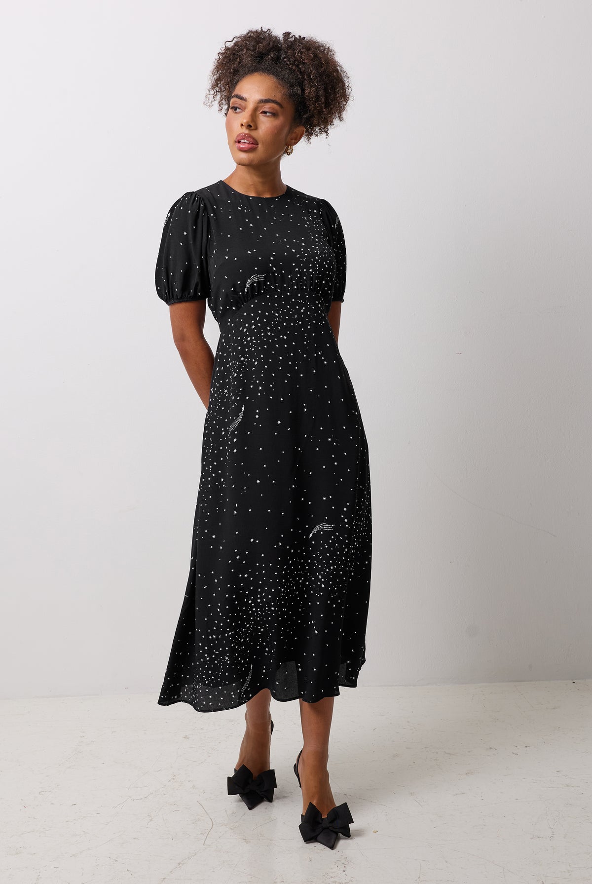 Scamp & Dude Black with White Written In The Stars Print Curved Waist Midi Dress | Model wearing a black midi dress with white star print and black bow detail high heels.
