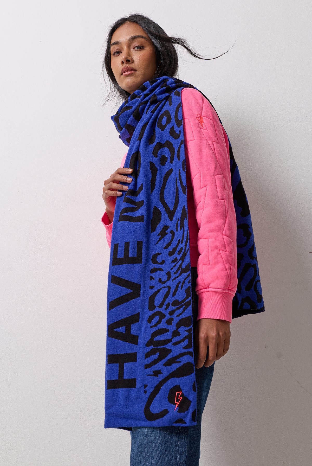 Scamp and Dude Navy with Black Slogan Jacquard Scarf | Model wearing a pair of blue jeans and a pink bomber jacket with a blue and black printed scarf.