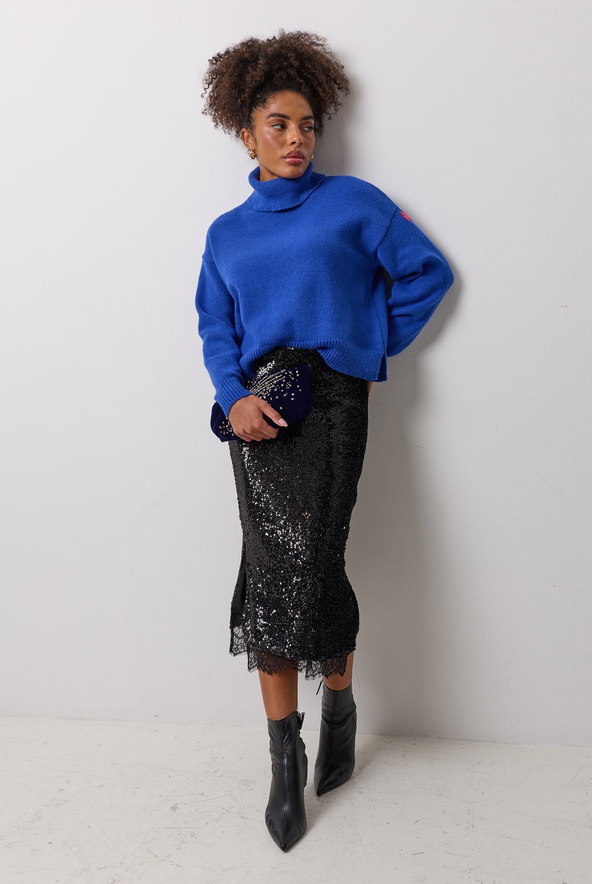 Scamp & Dude Black Sequin Lace Hem Midi Skirt | Model wearing a bright royal blue turtleneck sweater, a black sequined midi skirt, and black ankle boots.