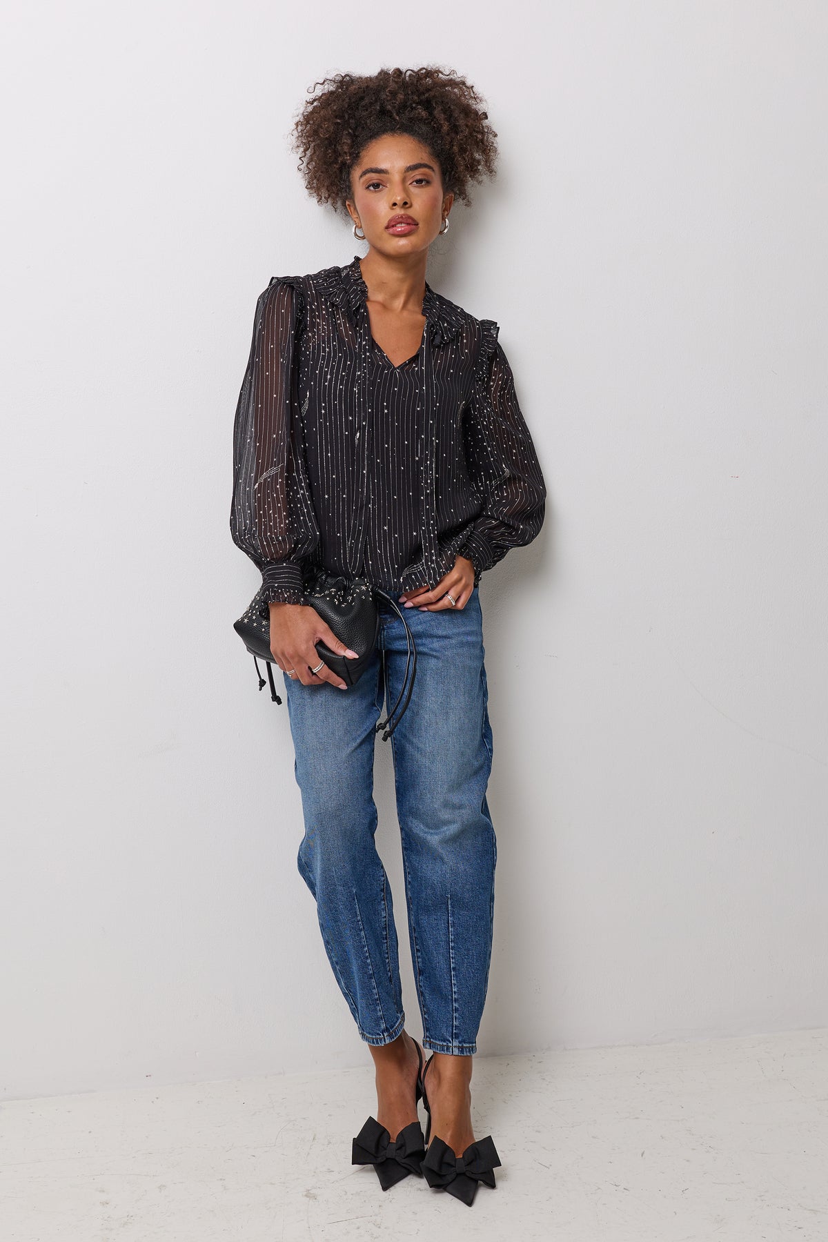 Scamp & Dude Black with Pewter Metallic Stripe and Star Blouse | Model wearing a pair of barrel leg denim jeans with a sheer metallic stripe and star print blouse. Worn with black bow heels and a small handbag.