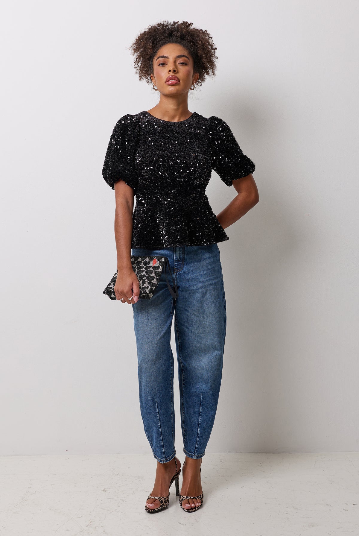 Scamp & Dude Black Sequin Peplum Top | Model wearing a black sequin peplum top with a pair of blue barrel jeans and a beaded clutch bag.