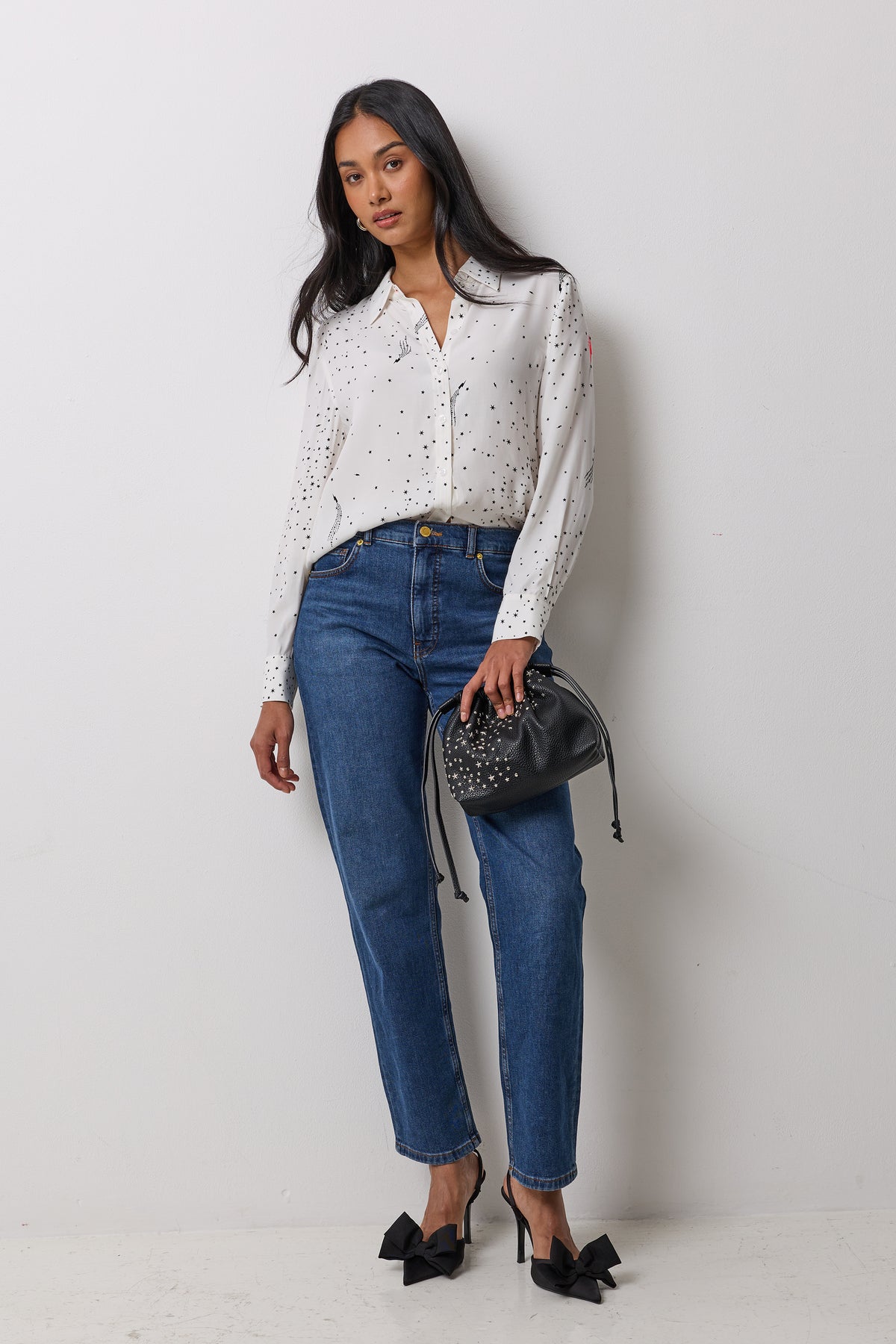 Scamp & Dude White with Black Written In The Stars Shirt | Model wearing a white long-sleeve button-up blouse with small black polka dots, paired with blue jeans, black bow heels, and holding a black studded bucket bag.