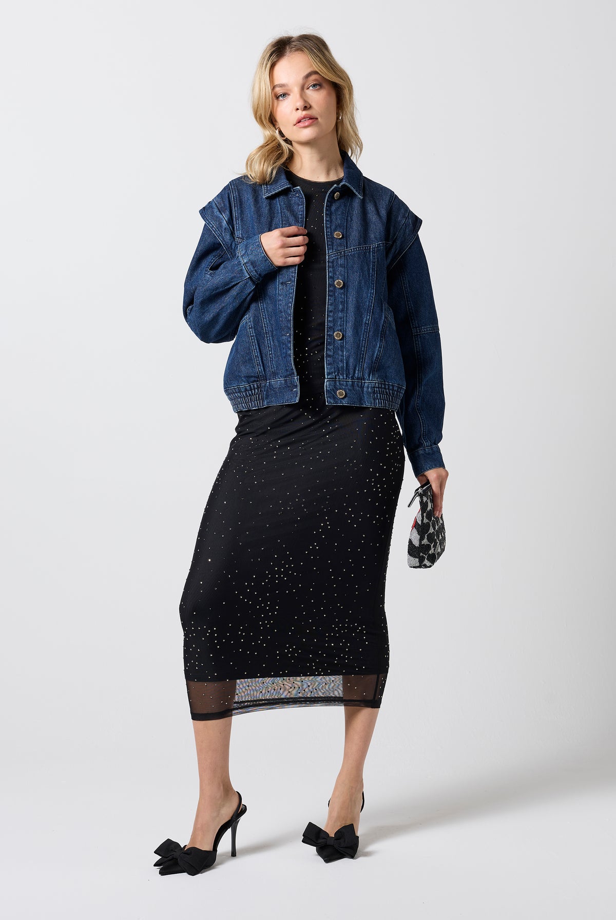 Scamp & Dude Mid Wash Seam Detail Denim Jacket | Model wearing a dark blue denim jacket over a black top, a black midi skirt with subtle silver speckles, and black pointed-toe heels. She is holding a small black-and-white beaded patterned clutch.