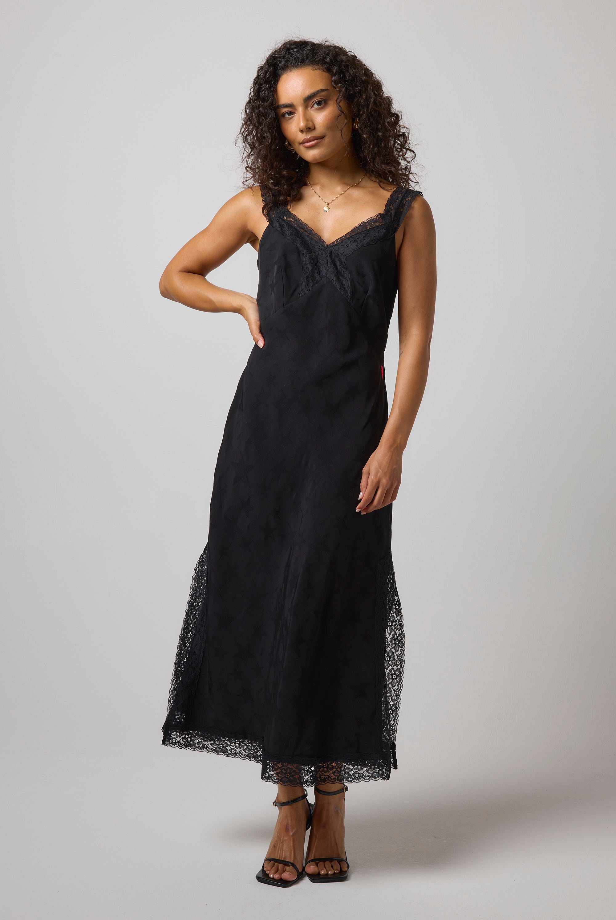 Scamp & Dude Black Star Jacquard Lace Detail Midi Slip Dress | Model wearing a sleeveless black midi dress with lace trim at the neckline and hem, paired with black strappy high-heels.