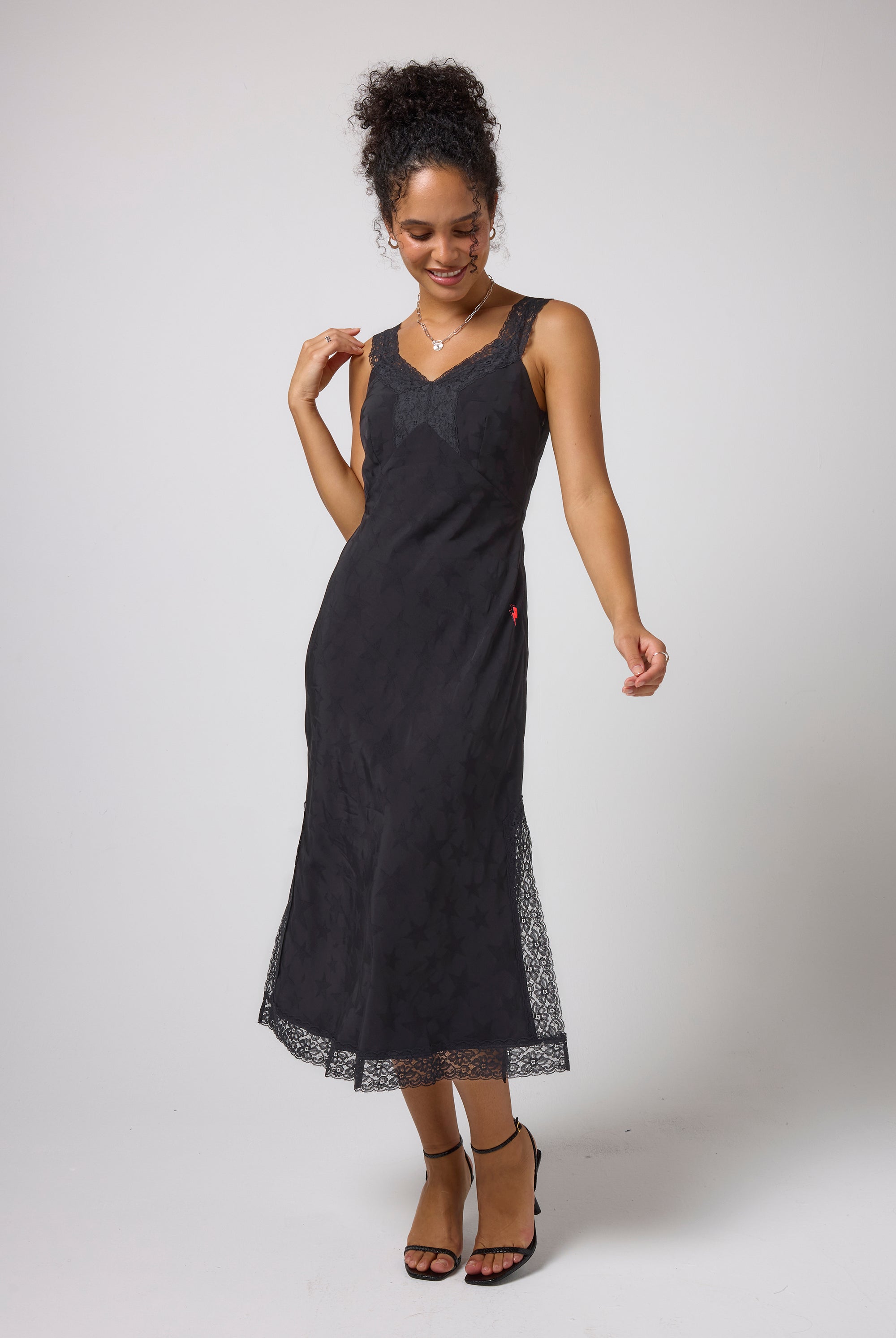 Scamp & Dude Black Star Jacquard Lace Detail Midi Slip Dress | Model wearing a sleeveless black midi dress with lace trim at the neckline and hem, paired with black strappy high-heels.