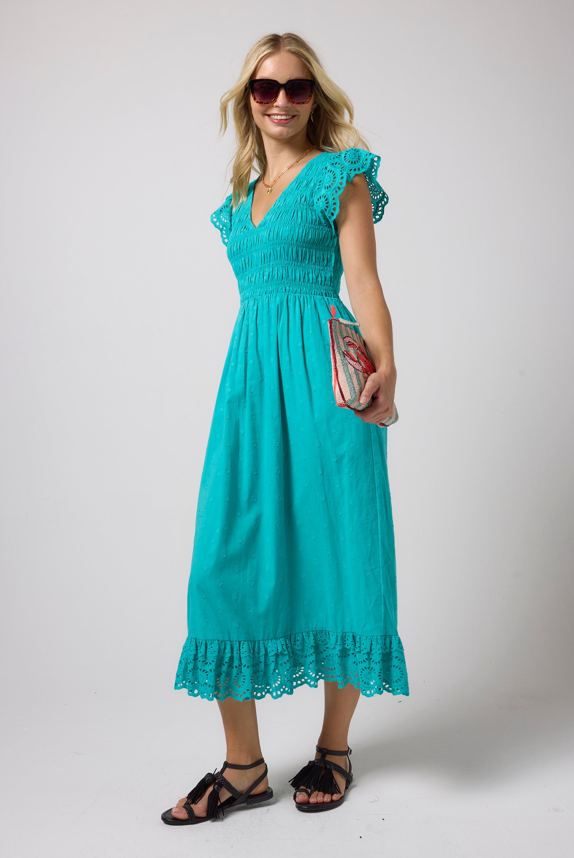 Scamp & Dude Turquoise Broderie Shirred Midi Dress | Model wearing a turquoise midi dress with broderie sleeves and a smocked bodice. Paired with black strappy sandals and tortoise shell sunglasses.