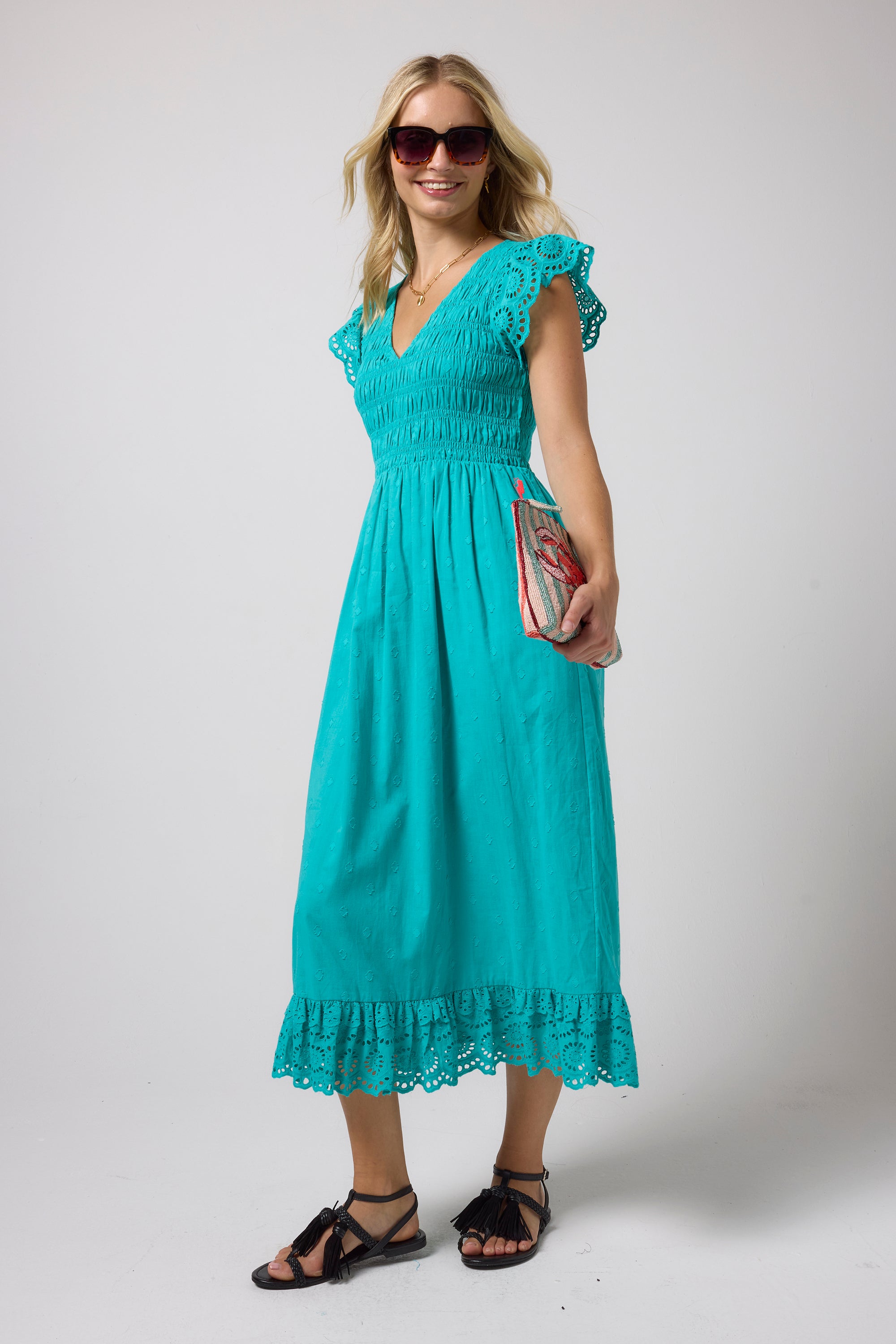 Scamp & Dude Turquoise Broderie Shirred Midi Dress | Model wearing a turquoise midi dress with broderie sleeves and a smocked bodice. Paired with black strappy sandals and tortoise shell sunglasses.