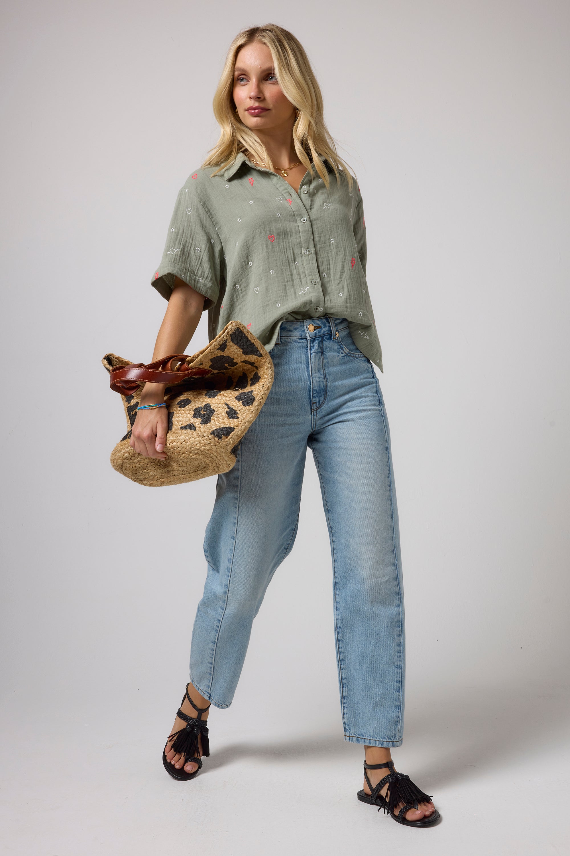 Scamp & Dude Khaki Cheesecloth Embroidered Shirt | Model wearing a light green short-sleeve button-up shirt, light-wash blue jeans, holding a woven straw bag with a leopard print.