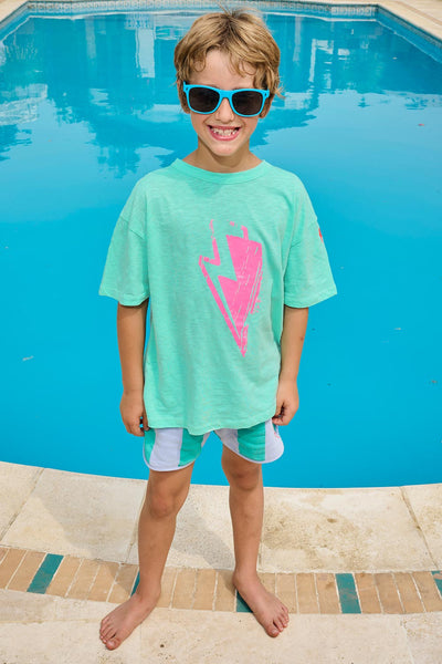 Scamp and Dude | Kids Mint with Neon Pink Bolt T-Shirt | Blonde boy wearing blue sunglasses and mint green t-shirt with pink lightening bolt in front of a swimming pool