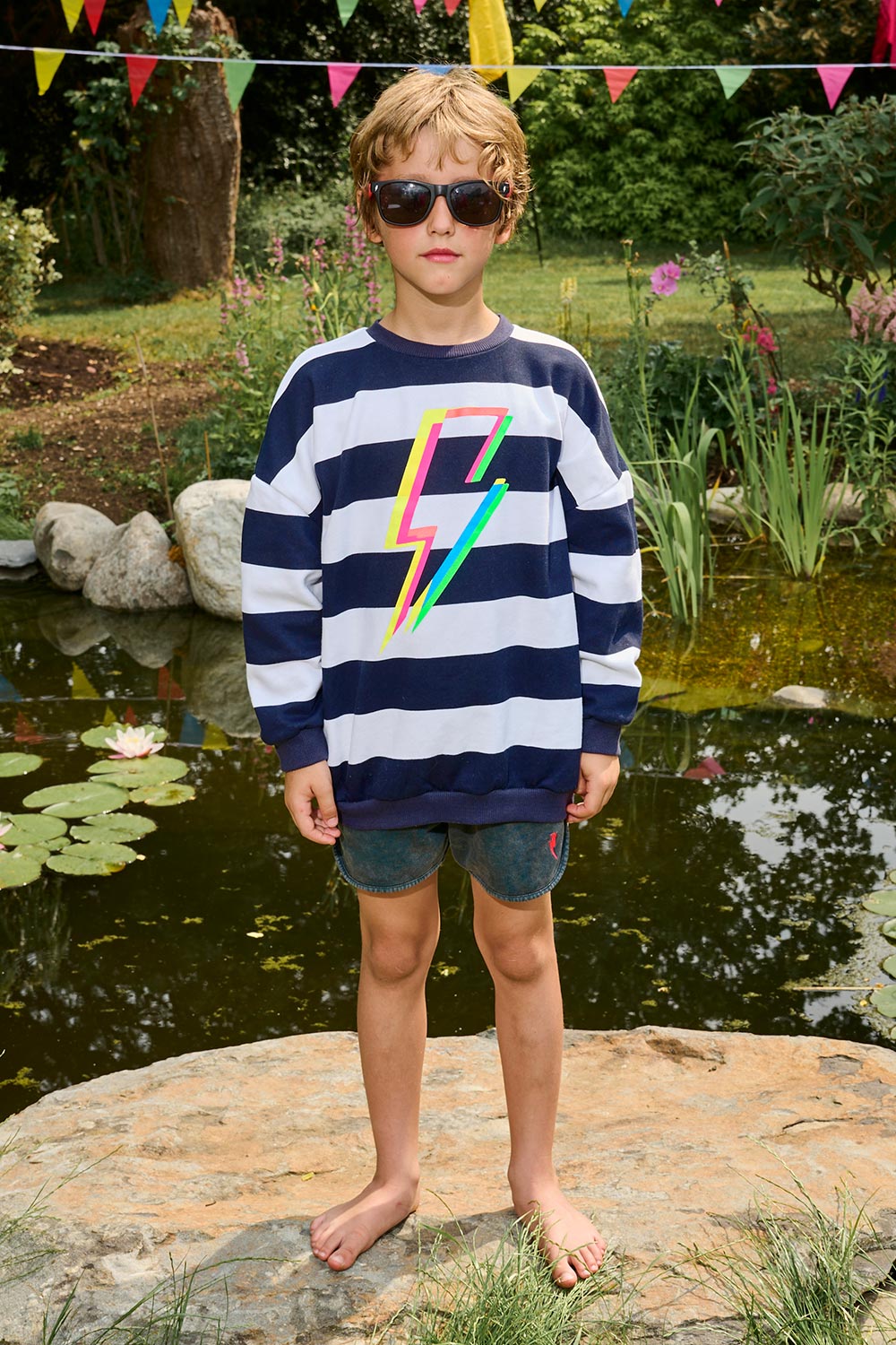 A boy wearing a navy with white stripe oversized sweatshirt with a multi-coloured neon lightning bolt on the front