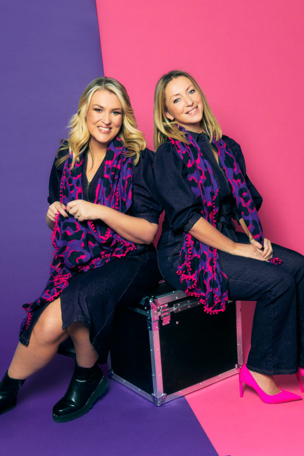 Scamp & Dude x Sara Davies Purple with Pink and Black Snow Leopard Charity Super Scarf | Two blonde women sat on a black box wearing navy dresses and purple and pink leopard print scarf with pink and purple background