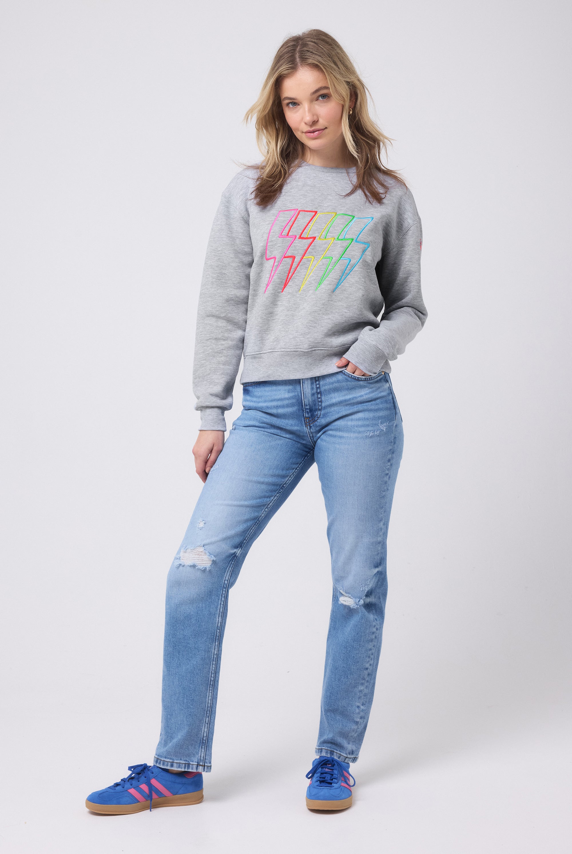 Scamp and Dude Grey Marl Multi Bolt Ultimate Sweatshirt | Model wearing a grey sweatshirt with multi coloured bolts detail. Worn with indigo distressed jeans and blue trainers.