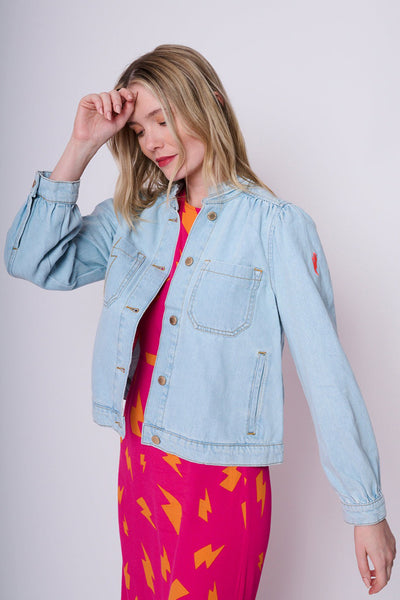 Scamp and Dude Pale Blue Wash Short Frill Collar Denim Jacket | Model with blonde hair holding hair with one hand wearing light wash denim jacket and bright pink patterned dress 