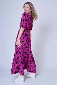 A blonde lady wearing a tiered skirt magenta maxi dress with black star and lightning bolt print and white shoes