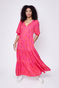 A curly-haired lady wearing a pink with orange zebra and lightning bolt print tie front tiered maxi dress with white trainers