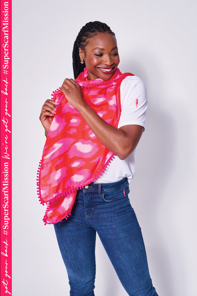 A lady wearing a red with hot pink and pale pink leopard ikat and lightning bolt print scarf with a pink pom pom trim